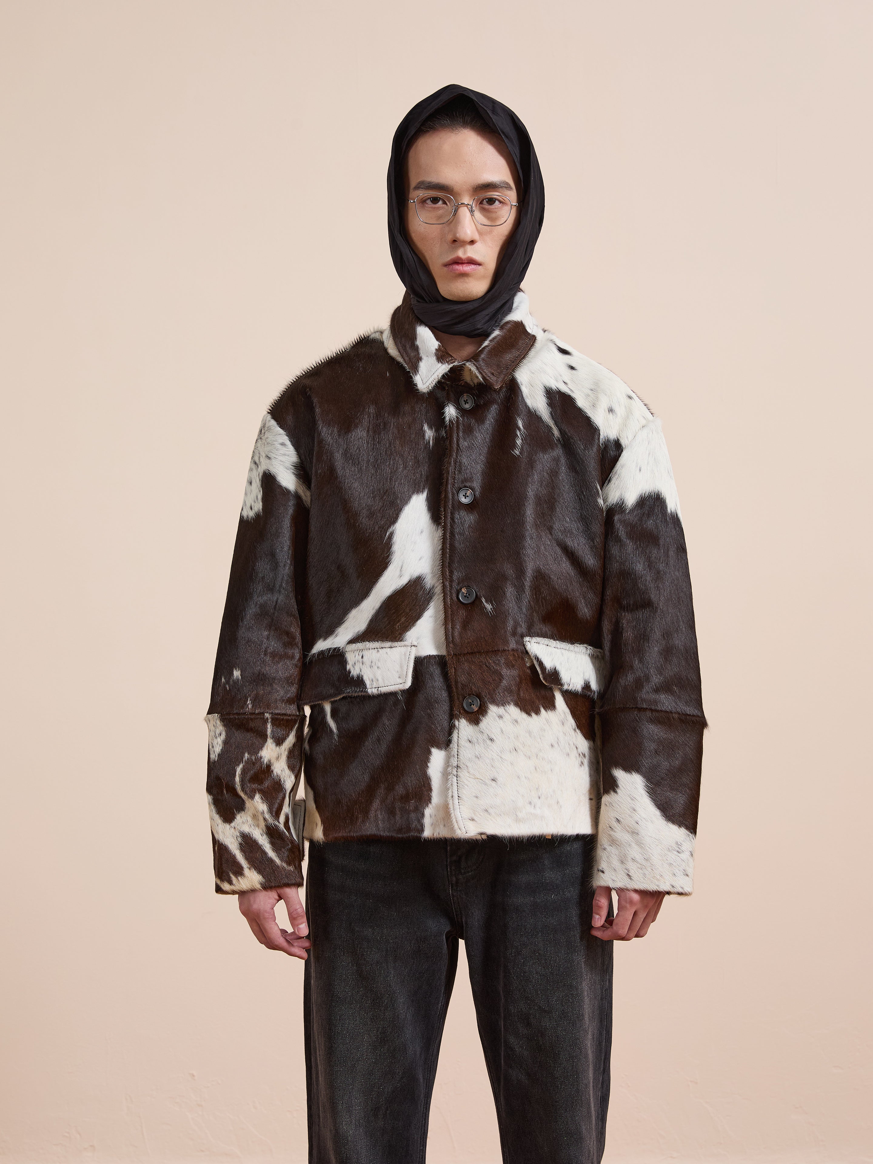 A person in glasses and a hood wears the FOUND Cowhide Prairie Utility Jacket, featuring a brown and white pattern, standing against a plain beige background to showcase its unique style.