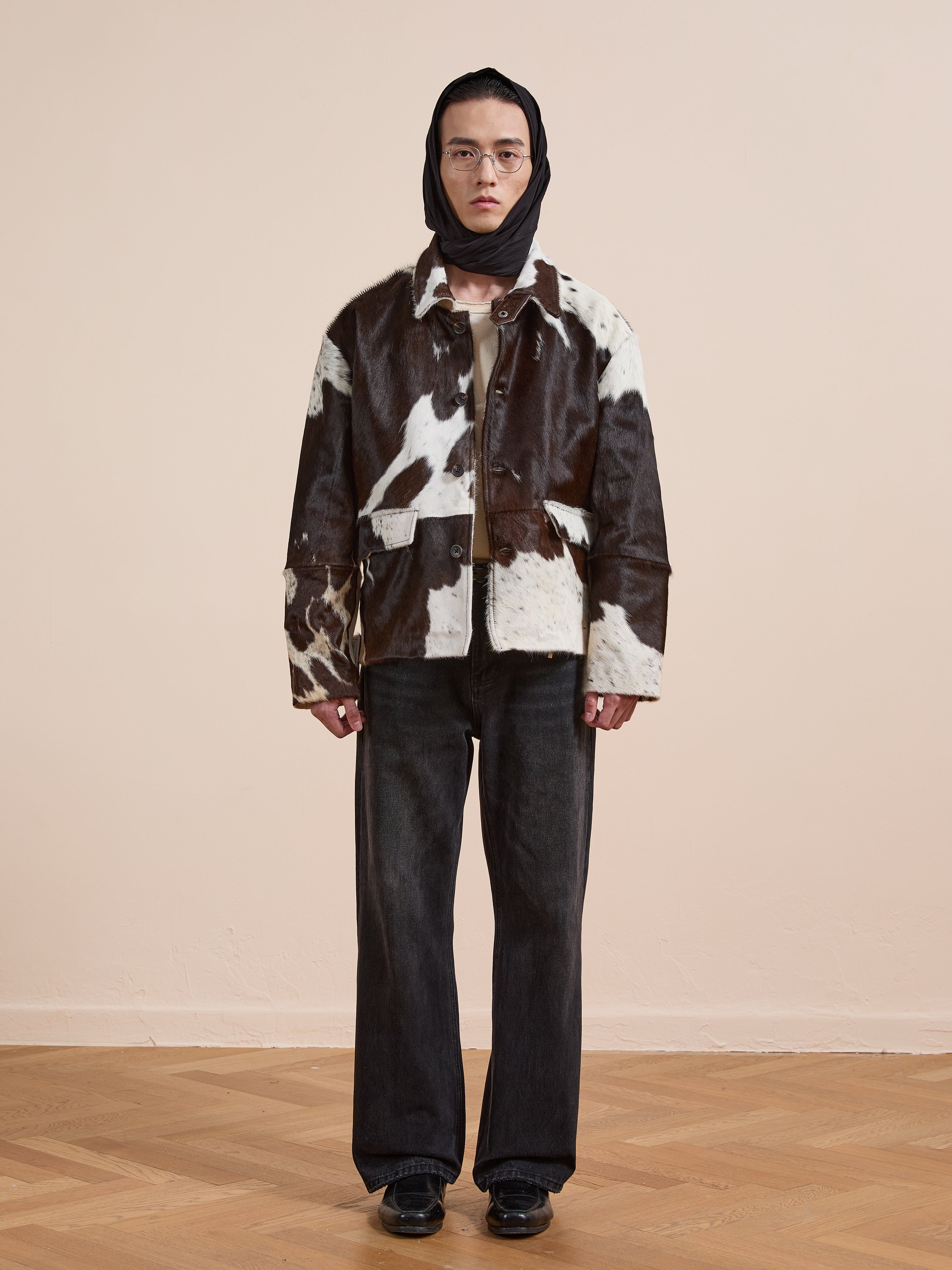 A person wearing a black headscarf, glasses, the FOUND Cowhide Prairie Utility Jacket, dark wide-leg jeans, and black shoes stands on a wooden floor against a plain beige wall.