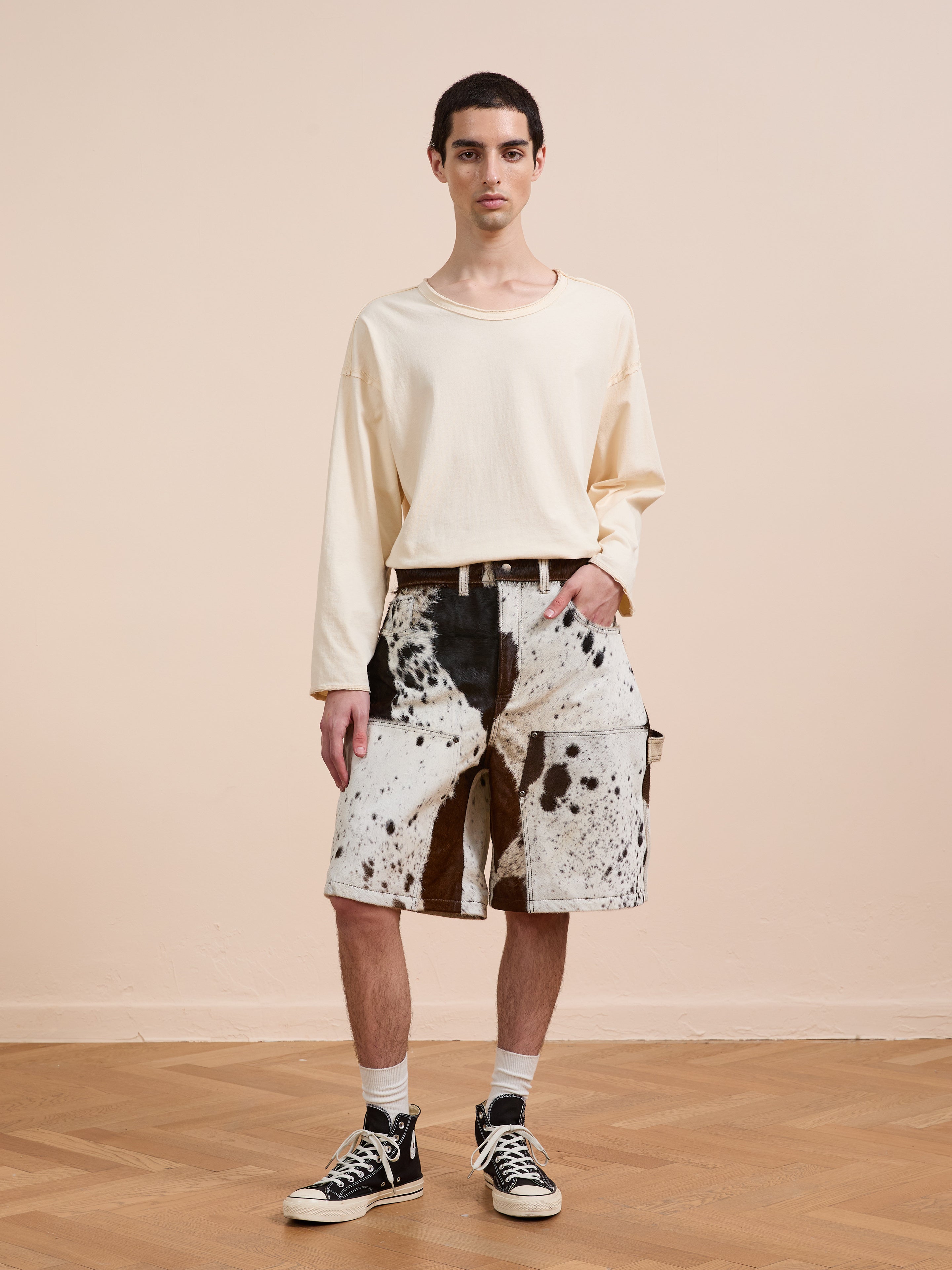 A person stands indoors on a wooden floor wearing a loose cream long-sleeve shirt, Found Cowhide Prairie Carpenter Shorts, white socks, and black sneakers against a plain beige background.