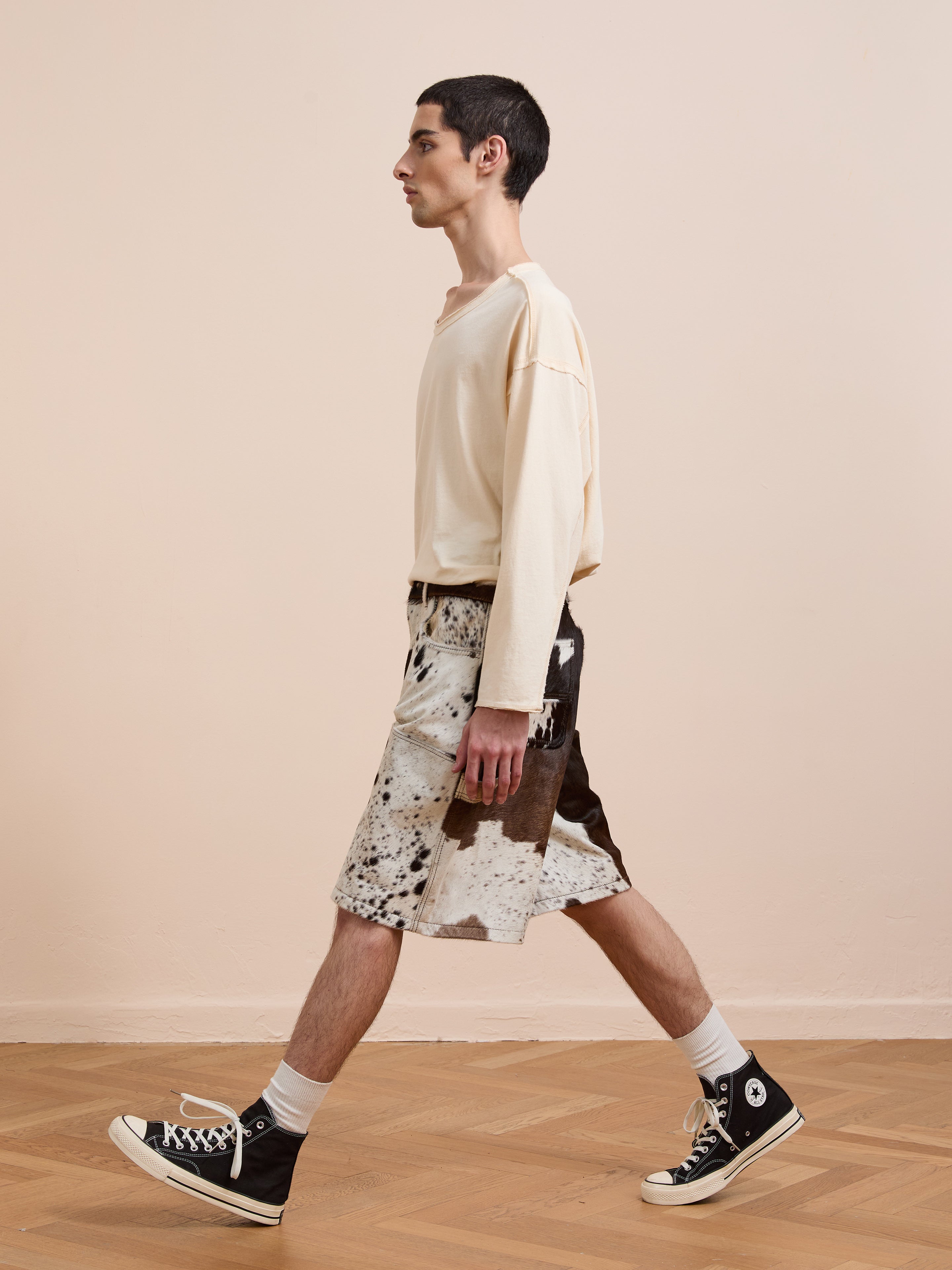 A person in a beige long-sleeve shirt, Found Cowhide Prairie Carpenter Shorts, white socks, and black high-top sneakers walks indoors on a wooden floor.