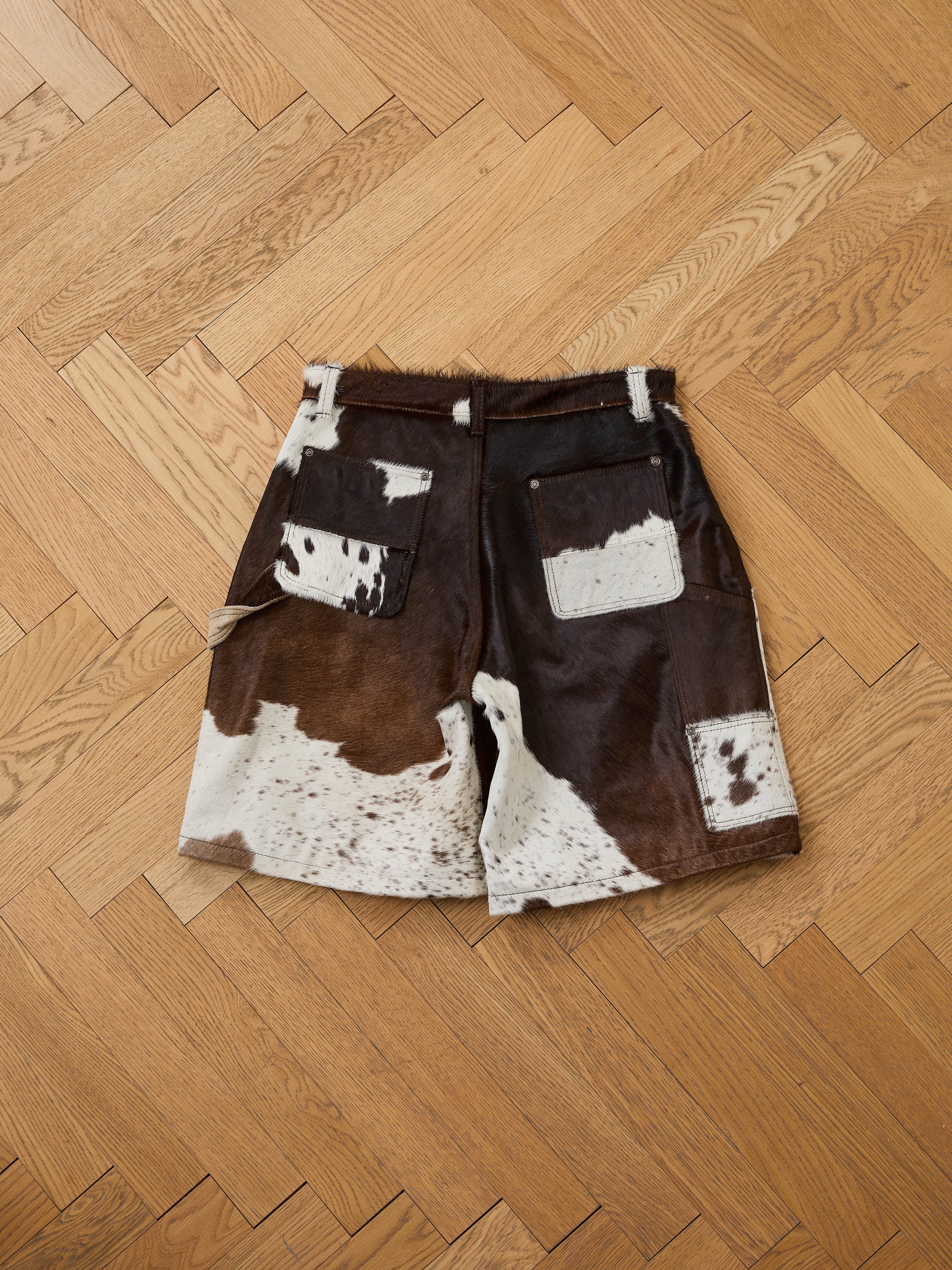 Found’s Cowhide Prairie Carpenter Shorts feature brown and white cowhide leather, a unisex relaxed fit, two back pockets, and are shown on a wooden herringbone floor.