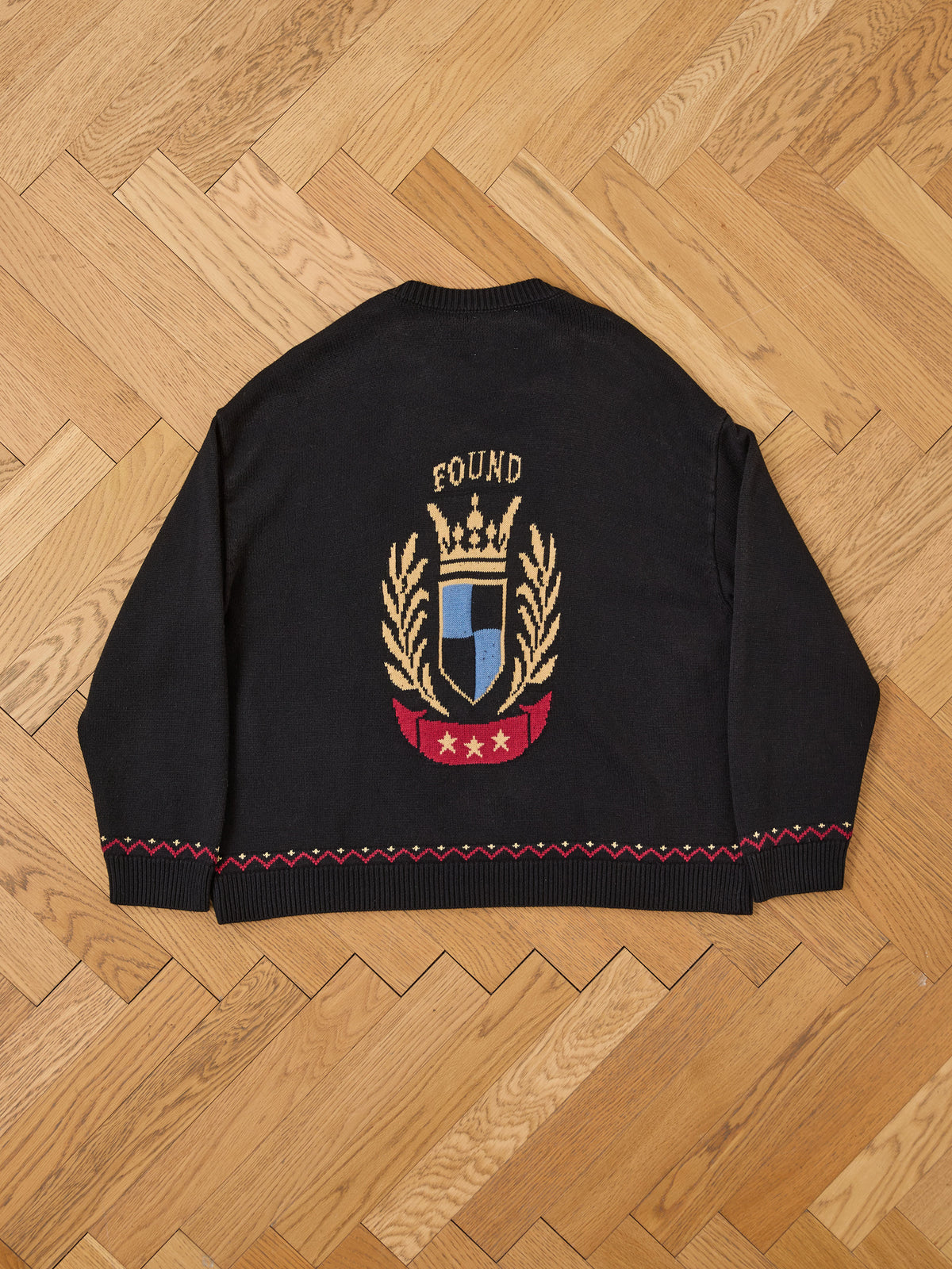 The Crest Sweater by FOUND features a gold laurel wreath, stars, shield, and “FOUND” on the back, styled like classic collegiate knitwear on a parquet floor.