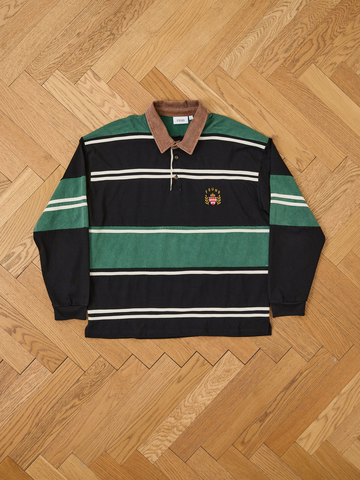 FOUND “Crestfield Rugby”—a long-sleeve unisex shirt with green/black stripes, brown collar, button placket, and embroidered logo—shown on a wooden herringbone floor.