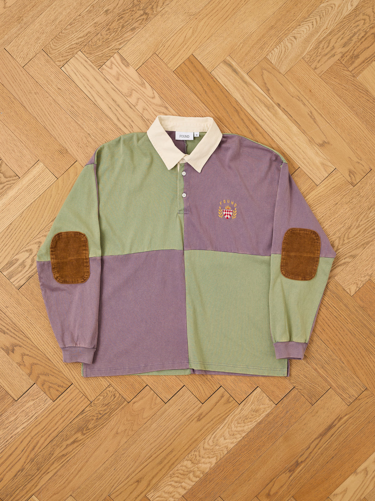 The FOUND Crestfield Rugby—a unisex rugby top with bold green and purple color-blocked panels, a beige collar, classic brown corduroy elbow patches, and an embroidered crest on the chest—is shown laid flat on a wooden floor.