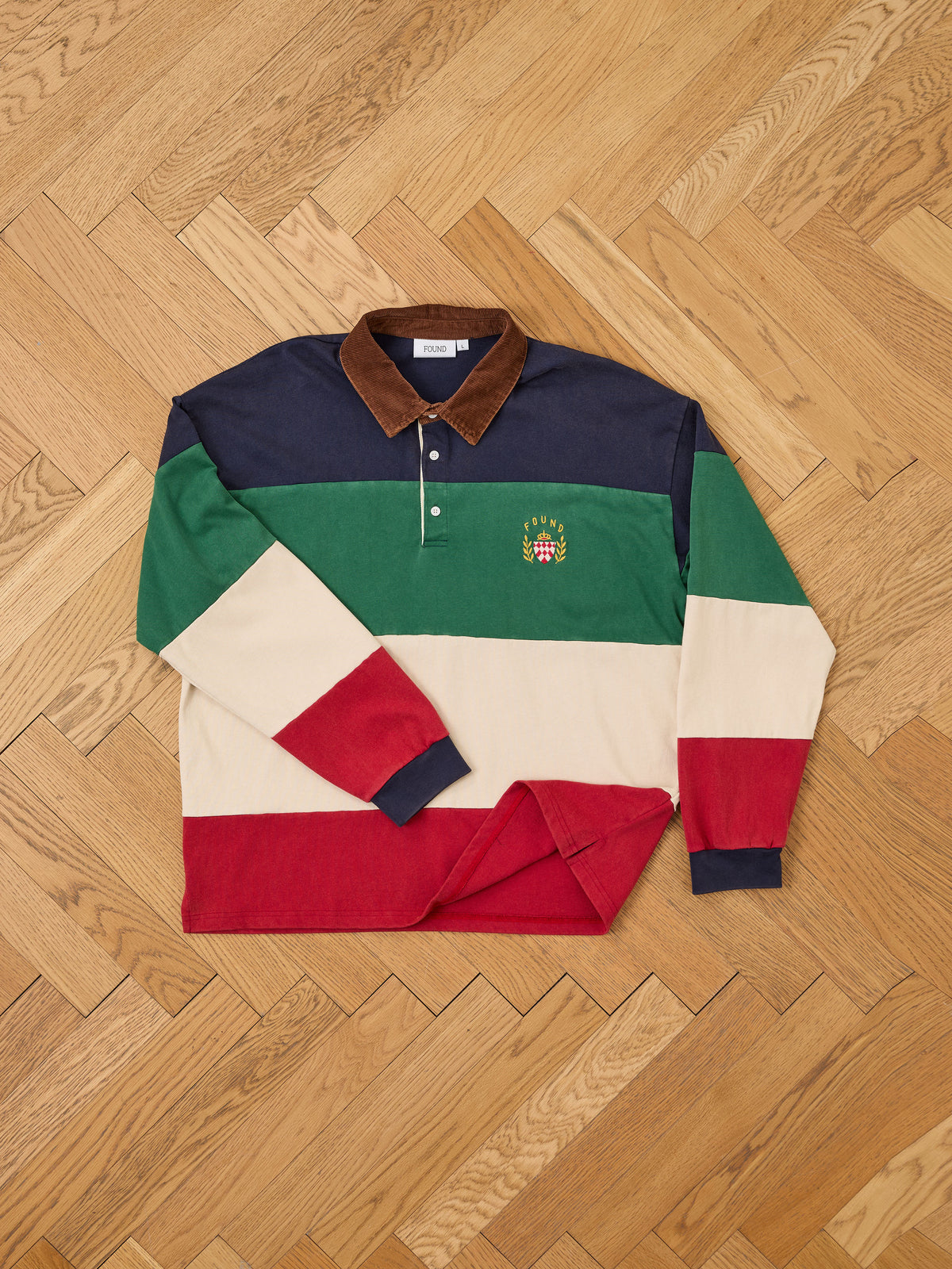 The FOUND Crestfield Rugby shirt, featuring navy, green, cream, and red horizontal stripes, a brown collar, and an embroidered crest on the chest, is displayed laid flat on a wooden herringbone floor.