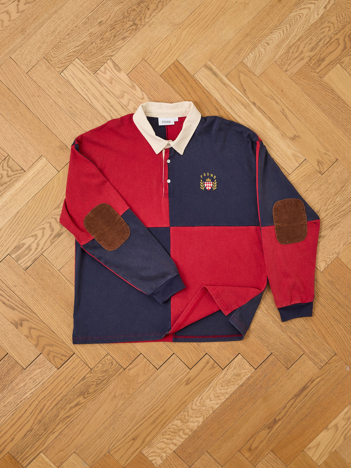 The FOUND Crestfield Rugby is a long-sleeve unisex relaxed fit shirt, showcased laid flat on a herringbone wood floor. It features bold red and navy blue color blocks, a tan collar, brown elbow patches, and an embroidered crest on the chest.