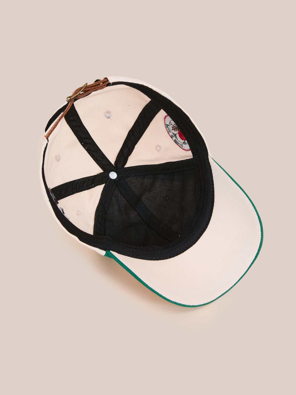 A light Crestline Racing Cap by Found, viewed from below, reveals black inner seams, a sweatband, and a metal buckle on its adjustable strap.