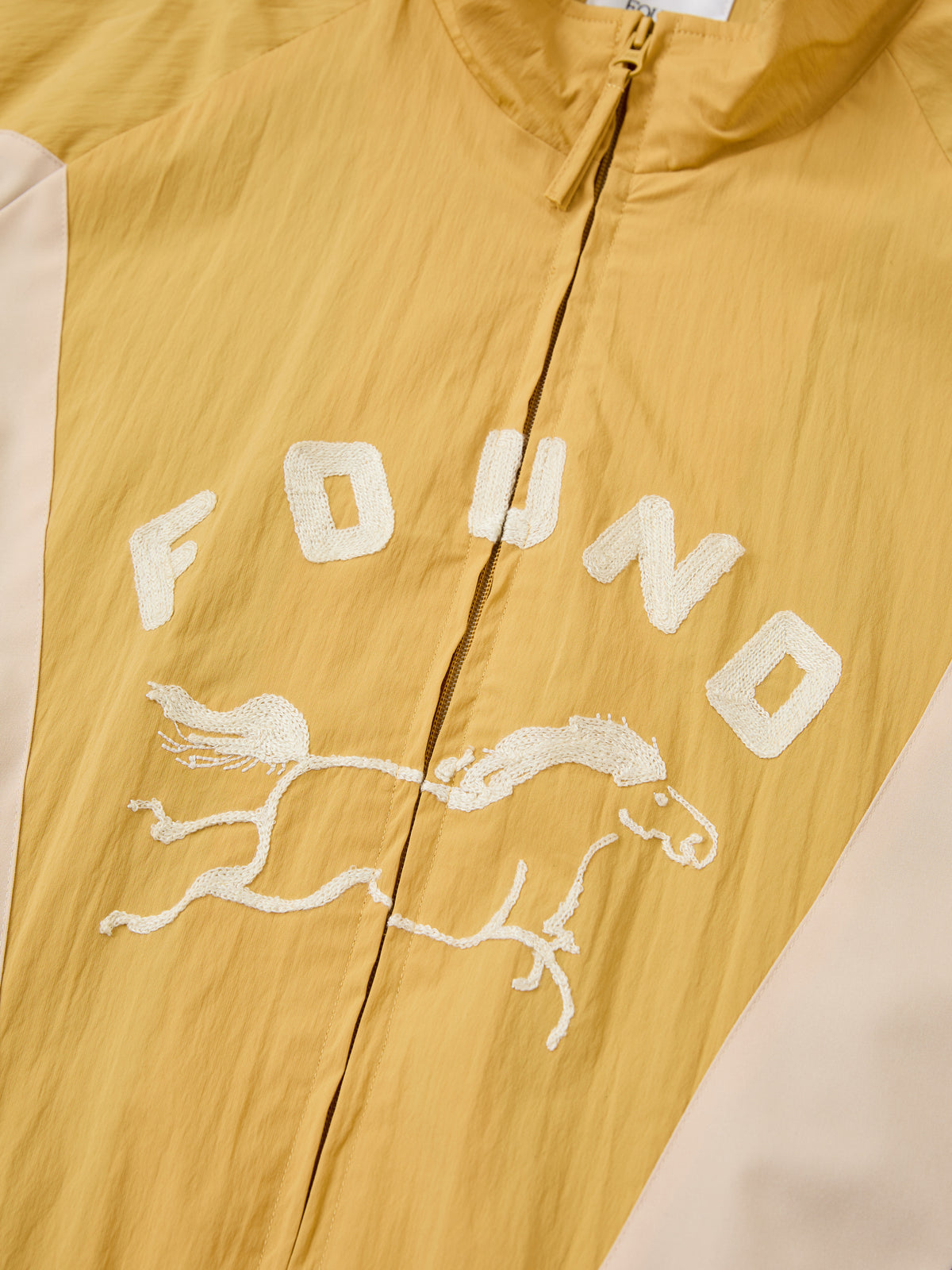 The Crestline Sport Zip Jacket by FOUND features yellow fabric, white panels, “FOUND” text, and an embroidered horse with rider on the front. Unisex and sporty.