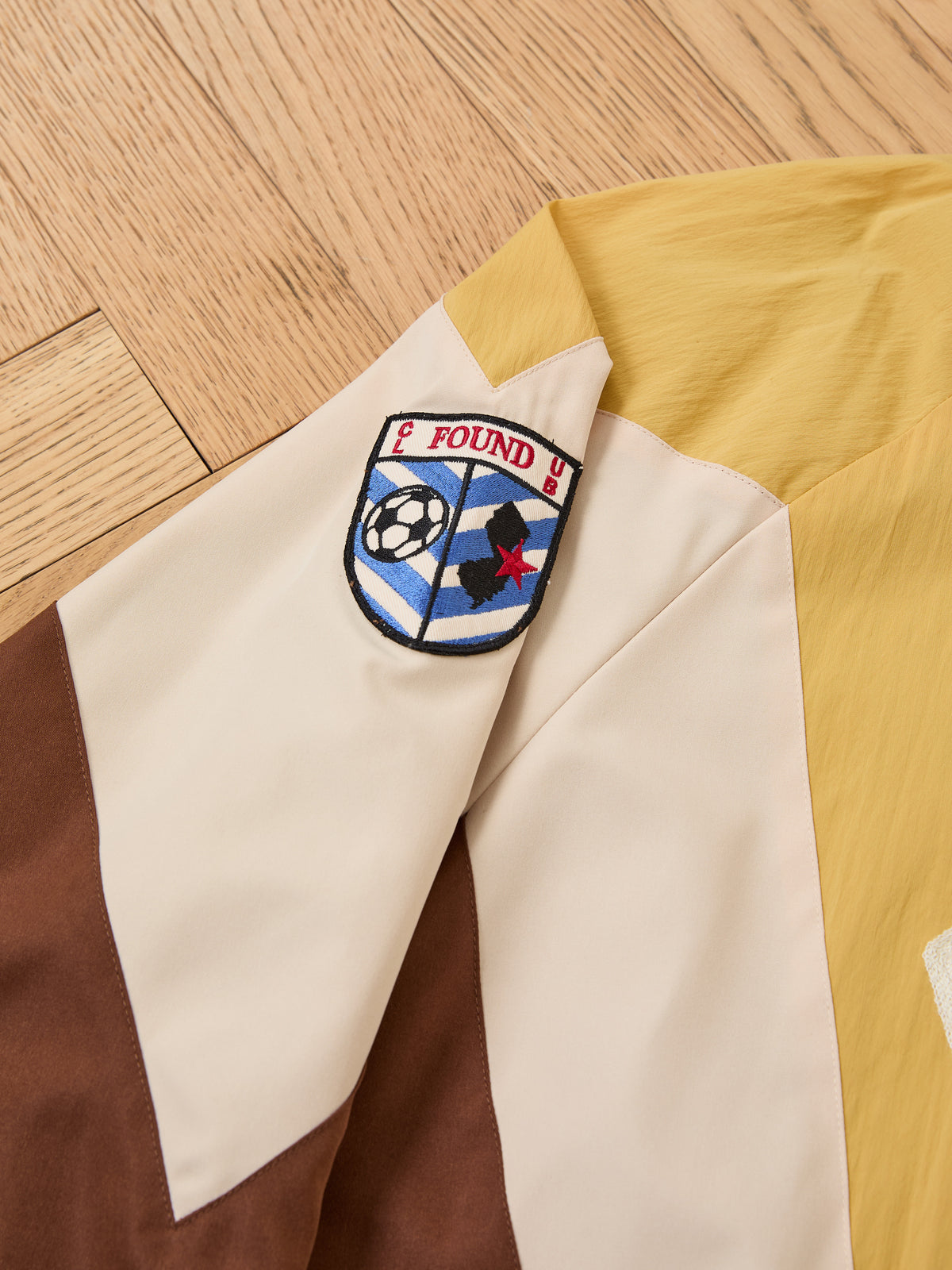 A beige, yellow, and brown Crestline Sport Zip Jacket by FOUND with a soccer ball patch and “FOUND U” on the sleeve is lying on a wooden floor.