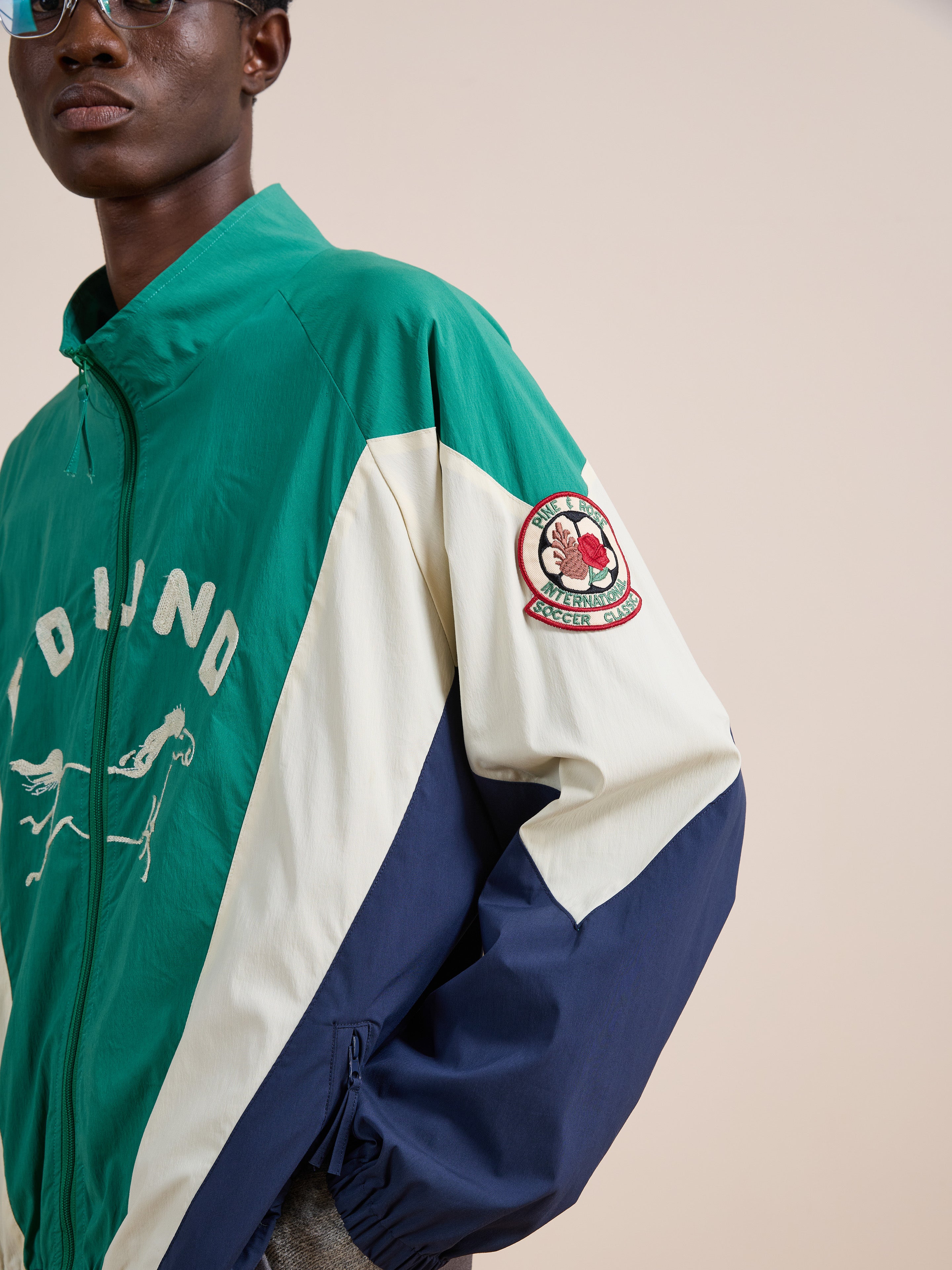A person wearing the FOUND Crestline Sport Zip Jacket in green, white, and navy, featuring a sleeve patch and chest graphic text for a classic vintage sportswear look.