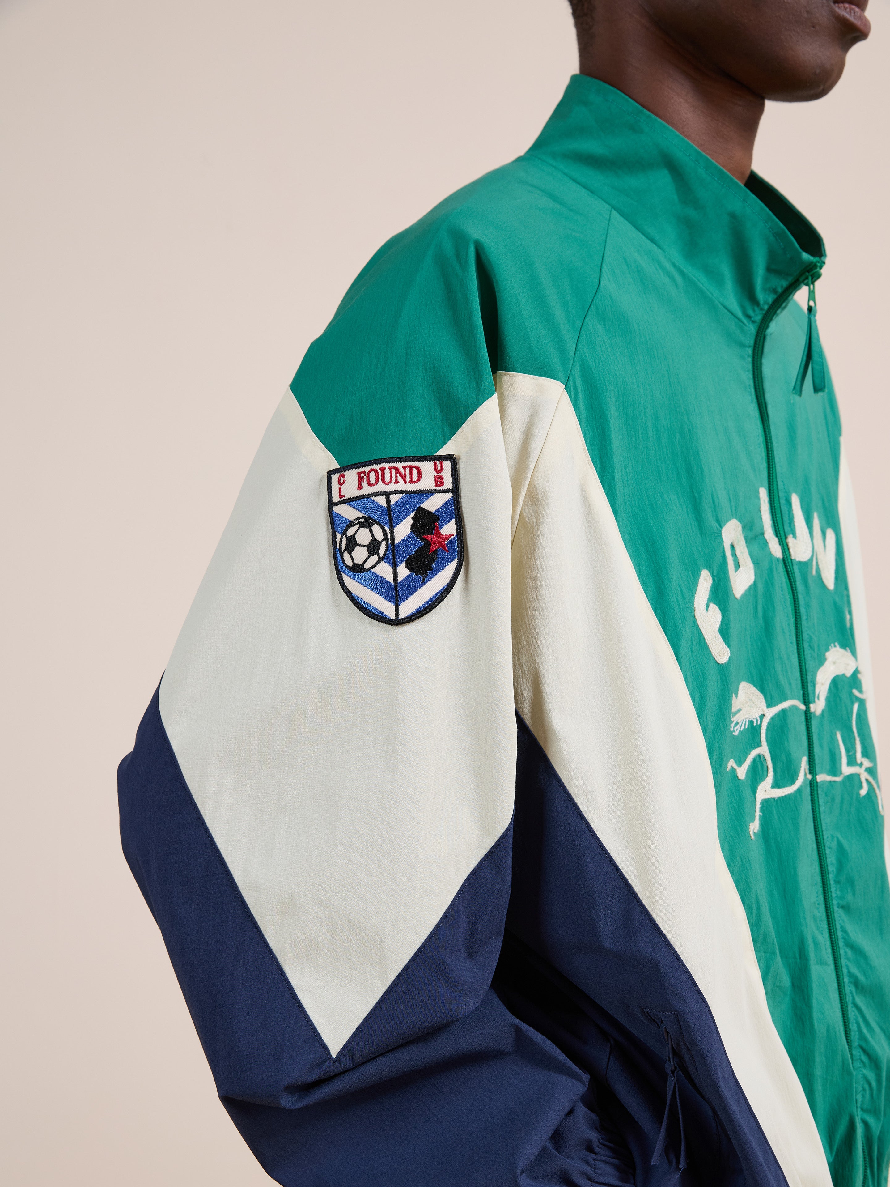 A person wears the FOUND Crestline Sport Zip Jacket in green, cream, and navy, featuring a sports-themed "FOUND" patch on the sleeve—a standout piece of vintage-inspired sportswear.