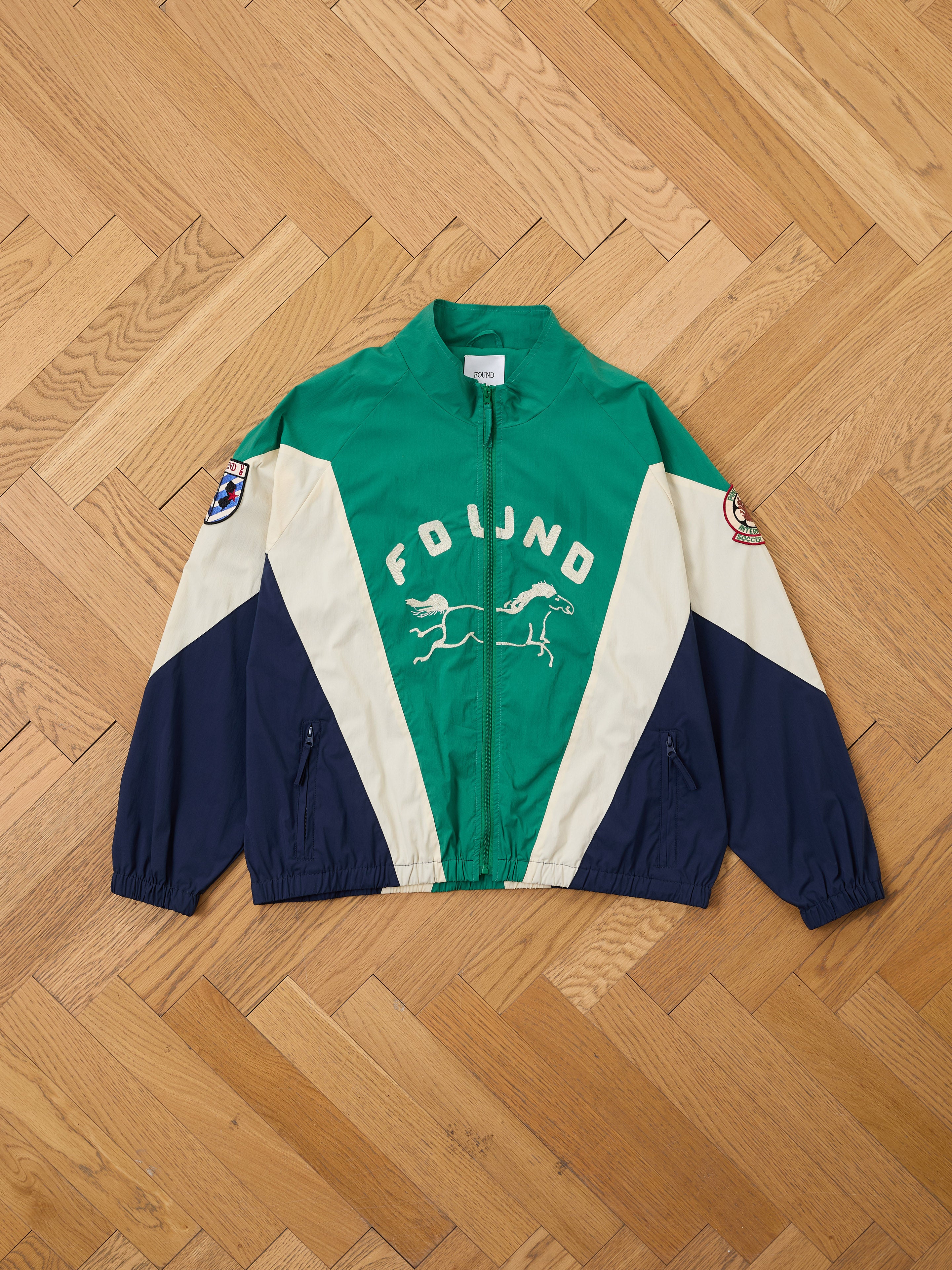 The Crestline Sport Zip Jacket by FOUND features green, navy, and cream panels, "FOUND" text, and two running horses on the front. Styled on a wooden parquet floor, this lightweight windbreaker channels vintage sportswear vibes.