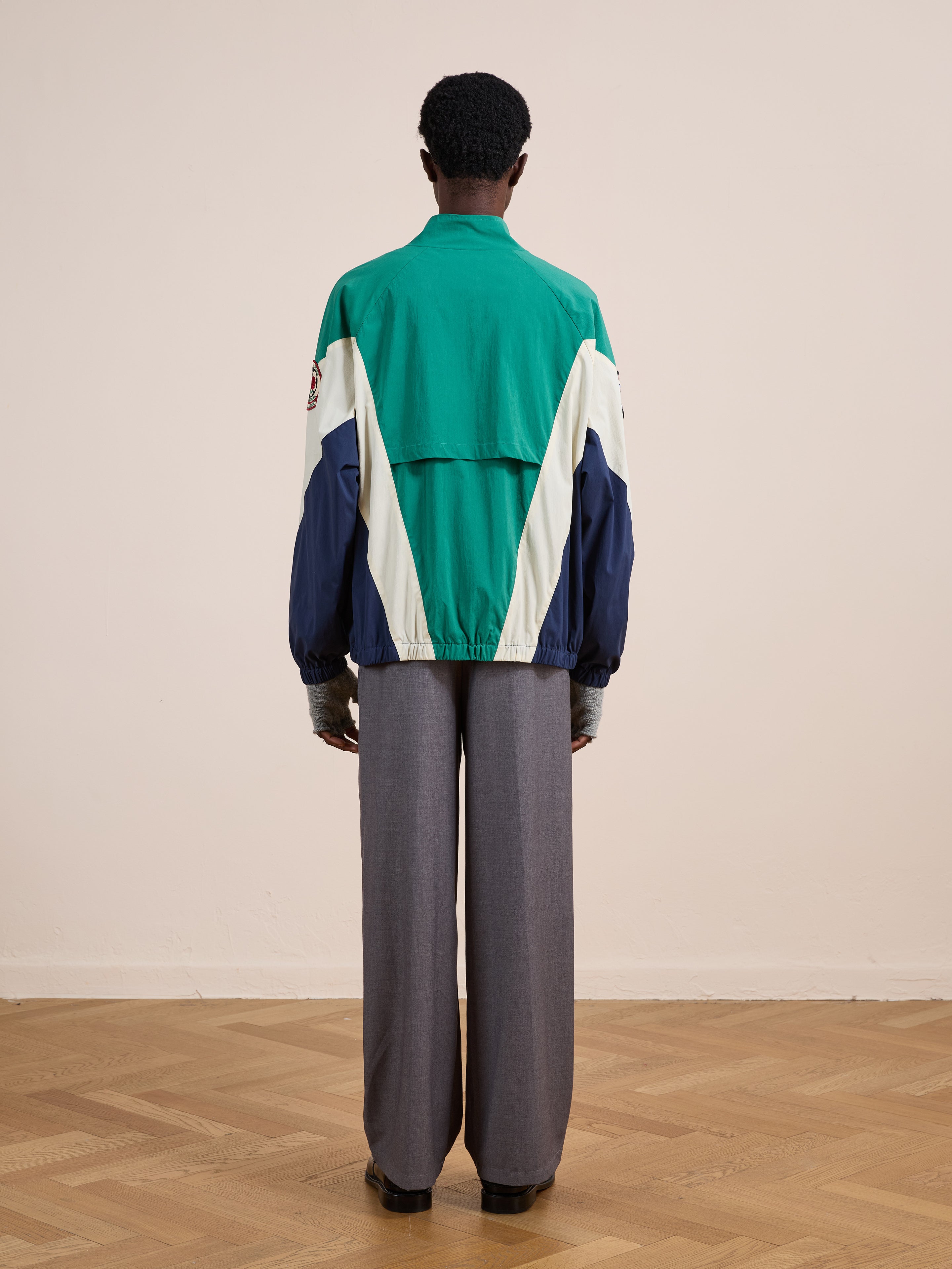 Indoors on a wooden floor, a person faces away from the camera wearing wide-leg gray pants and the FOUND Crestline Sport Zip Jacket in green, blue, and white with a classic vintage sportswear style.
