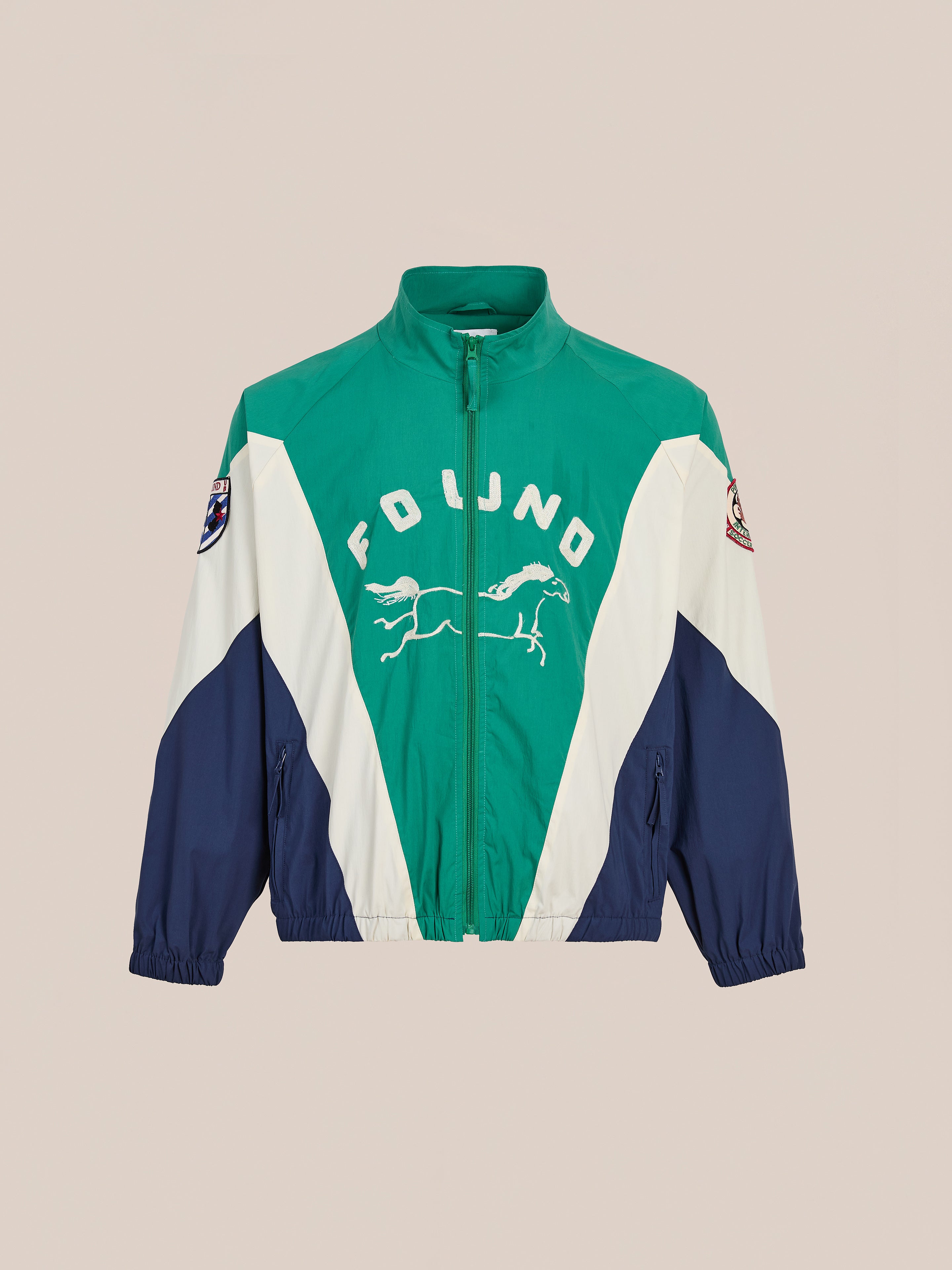 The Crestline Sport Zip Jacket by FOUND is a lightweight windbreaker with bold "FOUND" text, running horse graphics, sleeve patches, and retro color blocking in green, white, and navy—ideal for vintage sportswear fans.