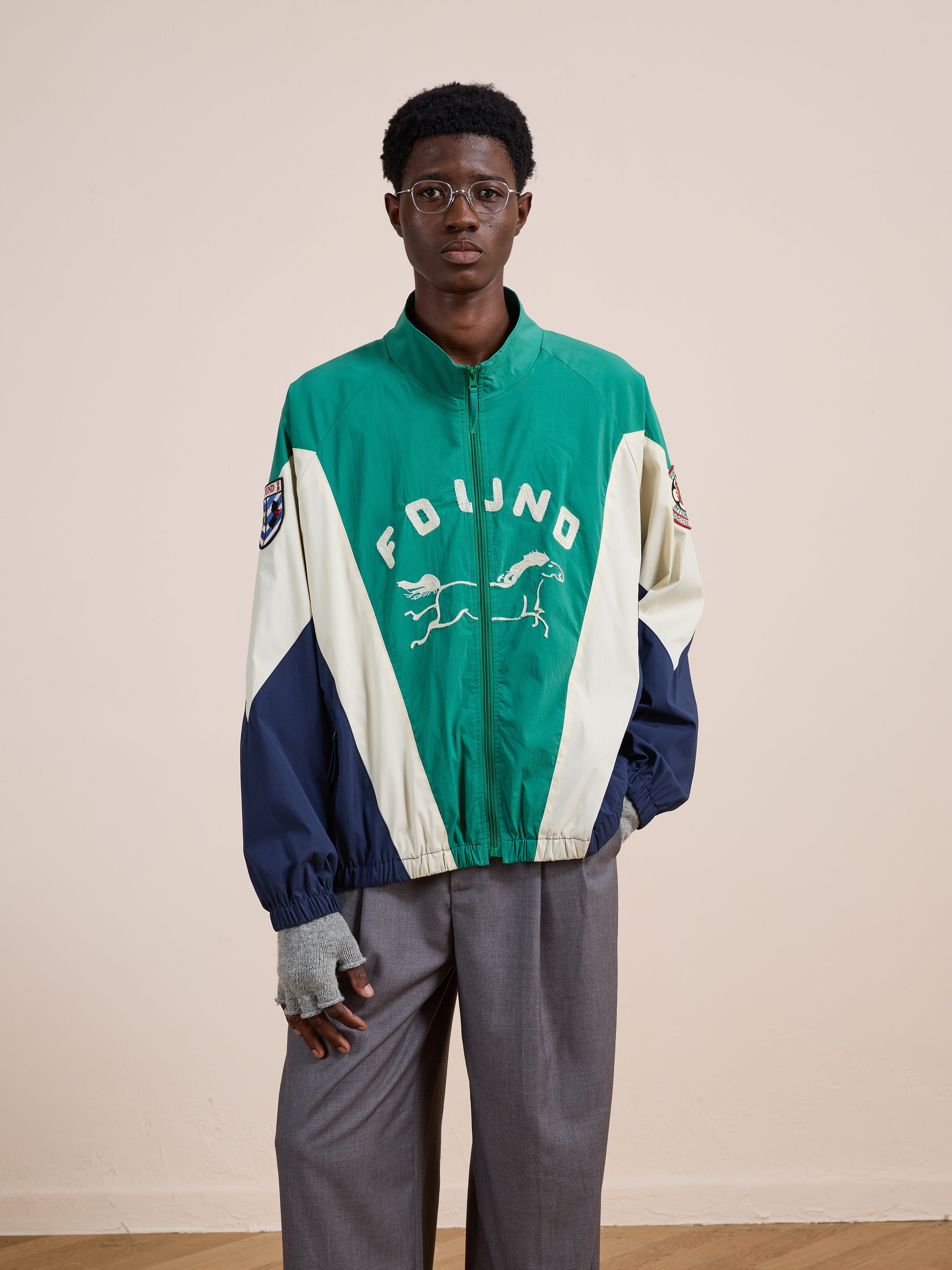 A person in glasses and gray pants models the FOUND Crestline Sport Zip Jacket in green and cream with a horse graphic, plus fingerless gloves, against a beige backdrop for a vintage sportswear vibe.