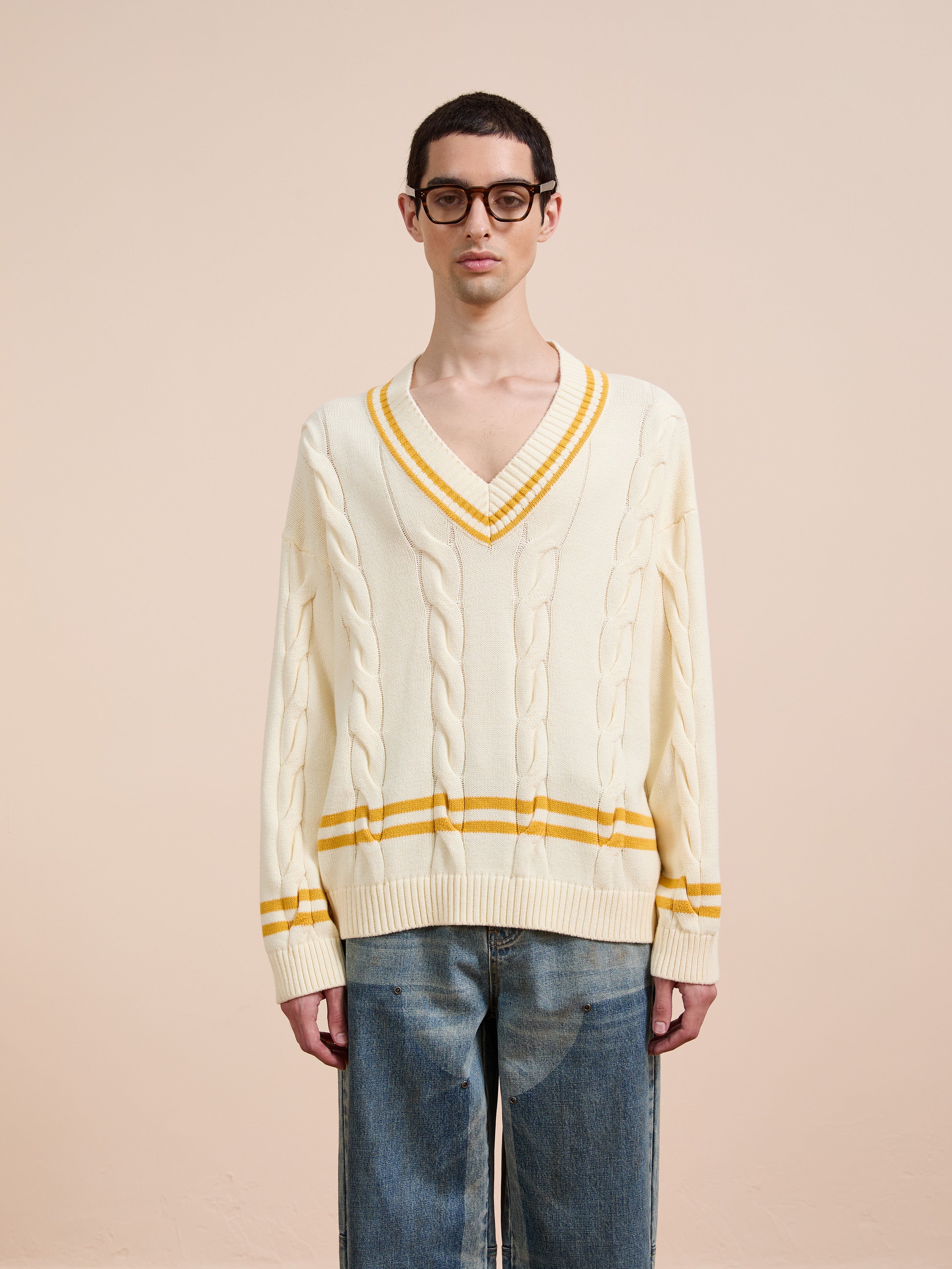 A person wearing glasses, a FOUND V-Neck Cricket Sweater with yellow stripes and blue jeans stands against a plain beige background.