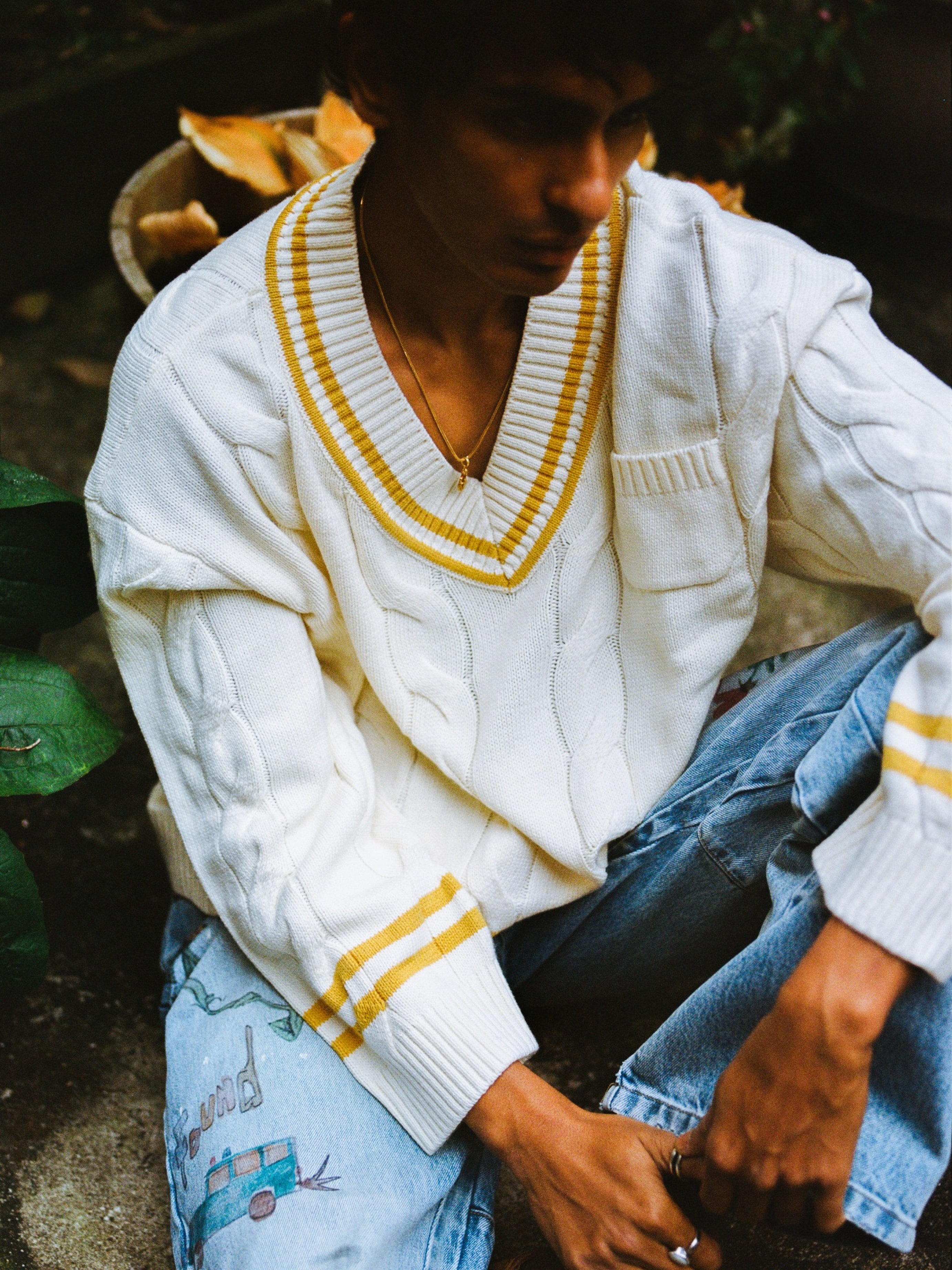 A person outdoors wears a FOUND V-Neck Cricket Sweater with yellow trim and blue jeans decorated with colorful embroidery.