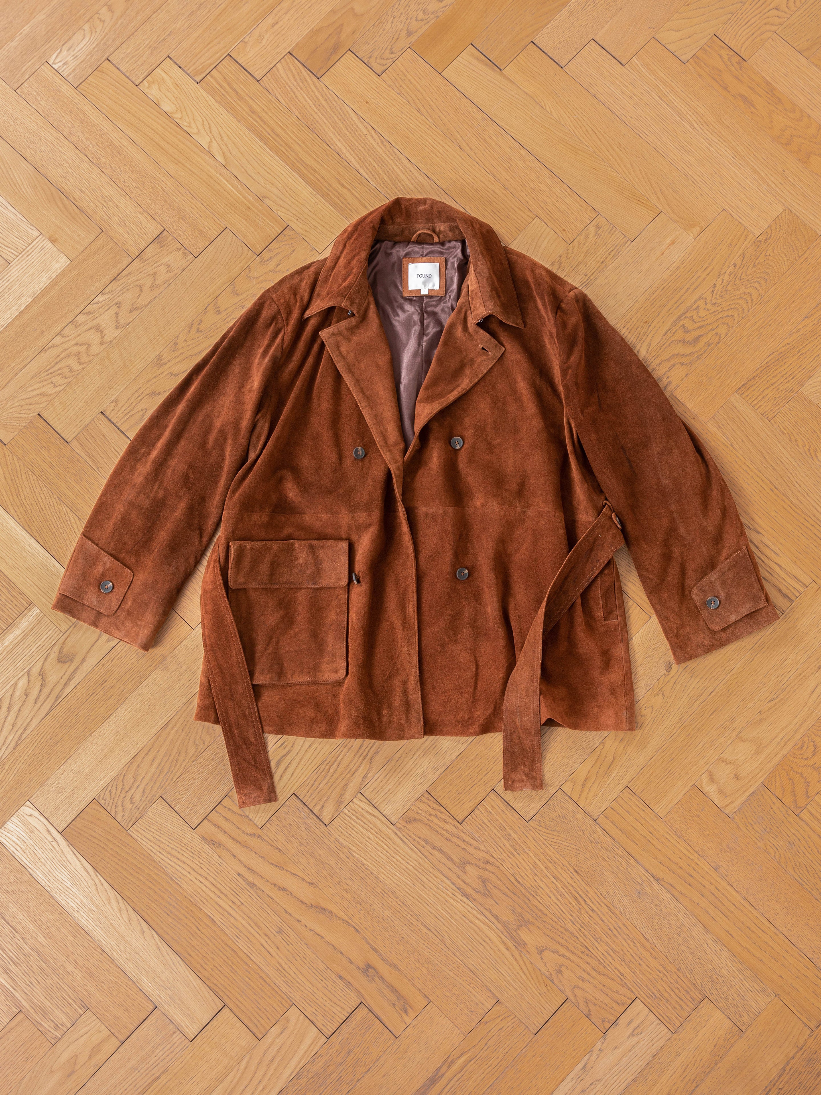 The FOUND Double Breasted Suede Leather Jacket, featuring wide lapels and a belt, is elegantly displayed on a chevron-patterned wooden floor.
