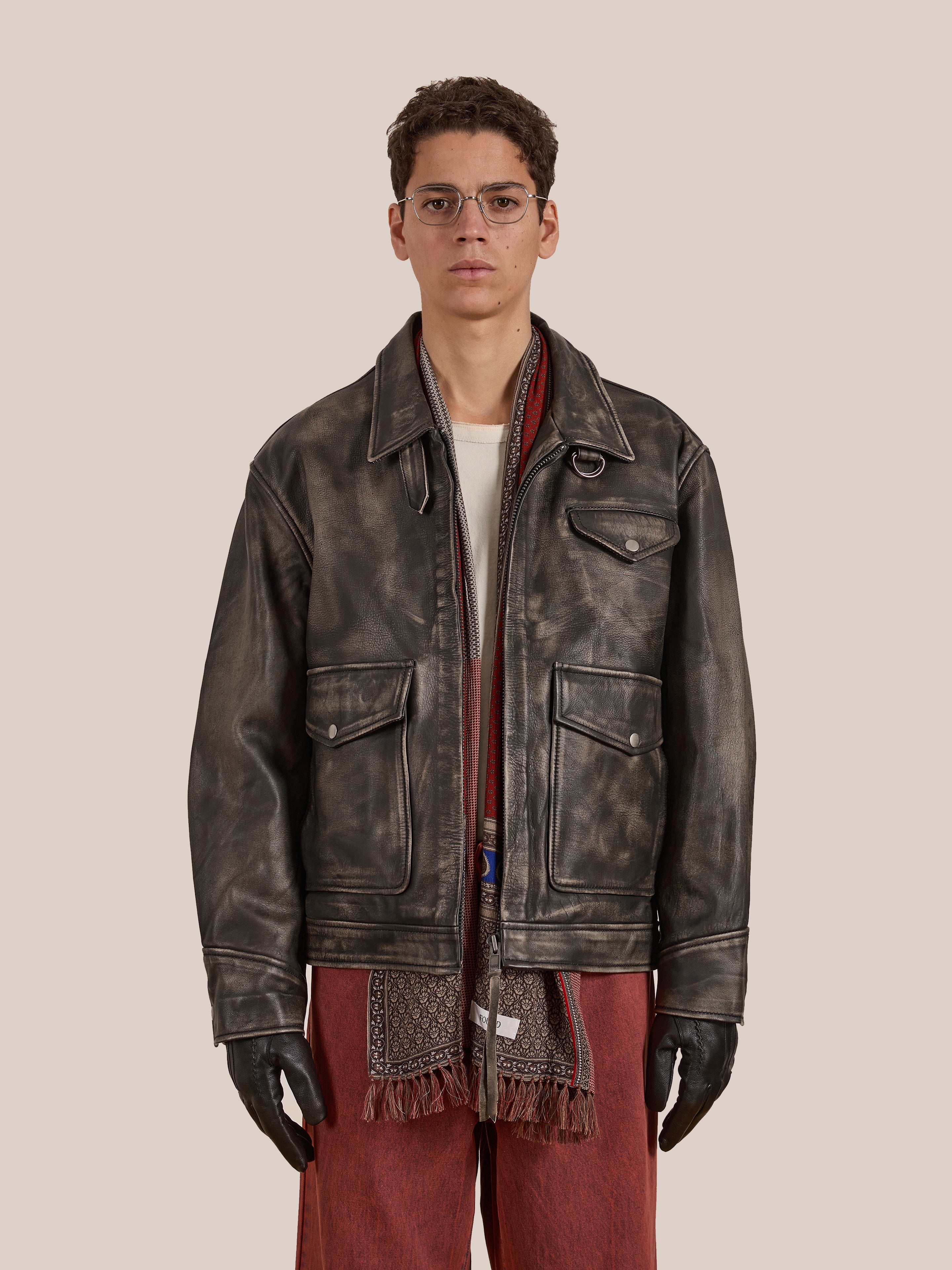 A person in Found's Distressed Leather Pocket Jacket, made from genuine cowhide, wearing a red scarf and brown pants with black gloves in a unisex relaxed fit set against a plain background.