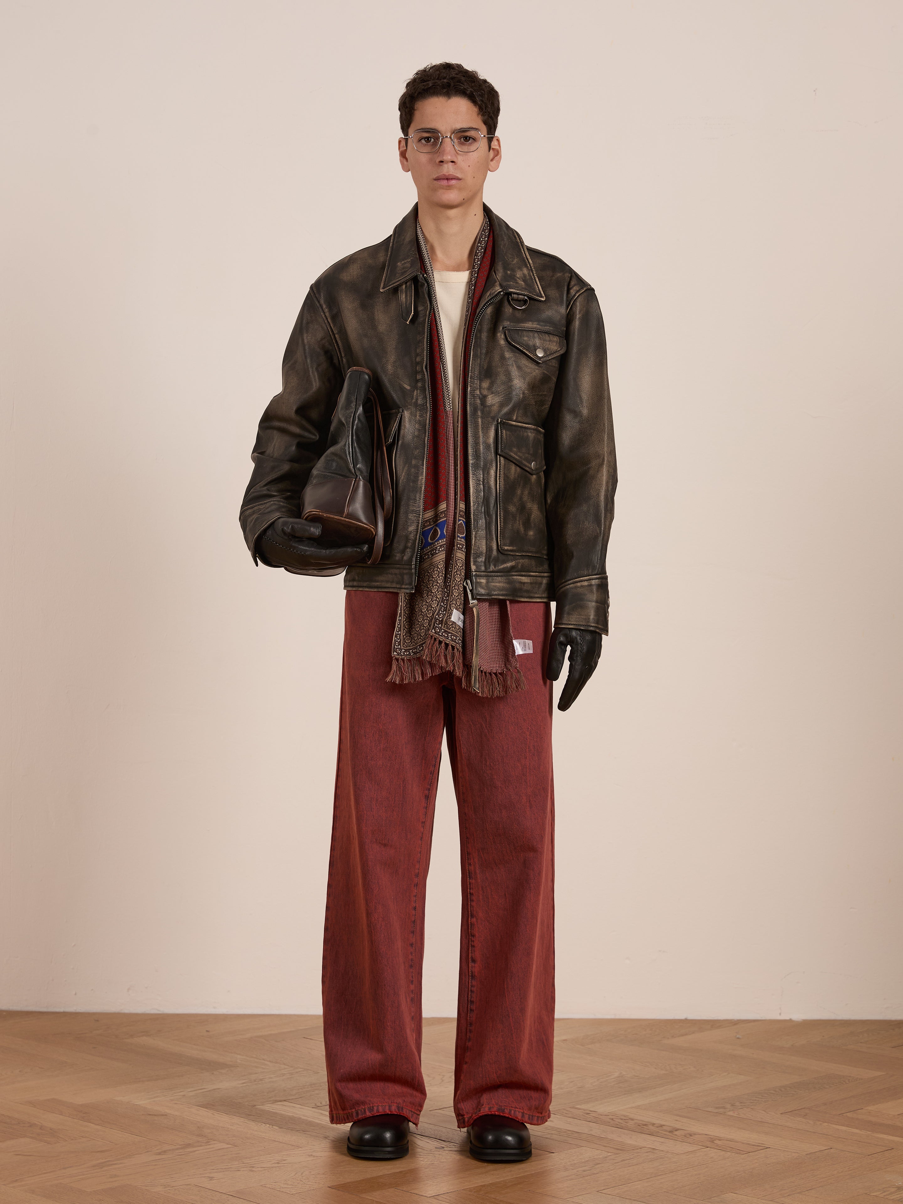 A person wearing a Found Distressed Leather Pocket Jacket, red pants, glasses, and gloves is holding a bag, standing on a wooden floor against a plain wall.