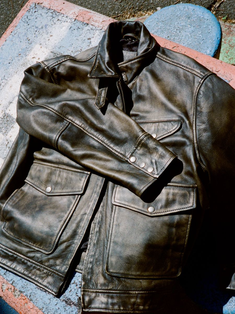 The Found Distressed Leather Pocket Jacket, a unisex relaxed fit black leather jacket with front pockets and snap buttons, is displayed on a textured, multicolored concrete surface.