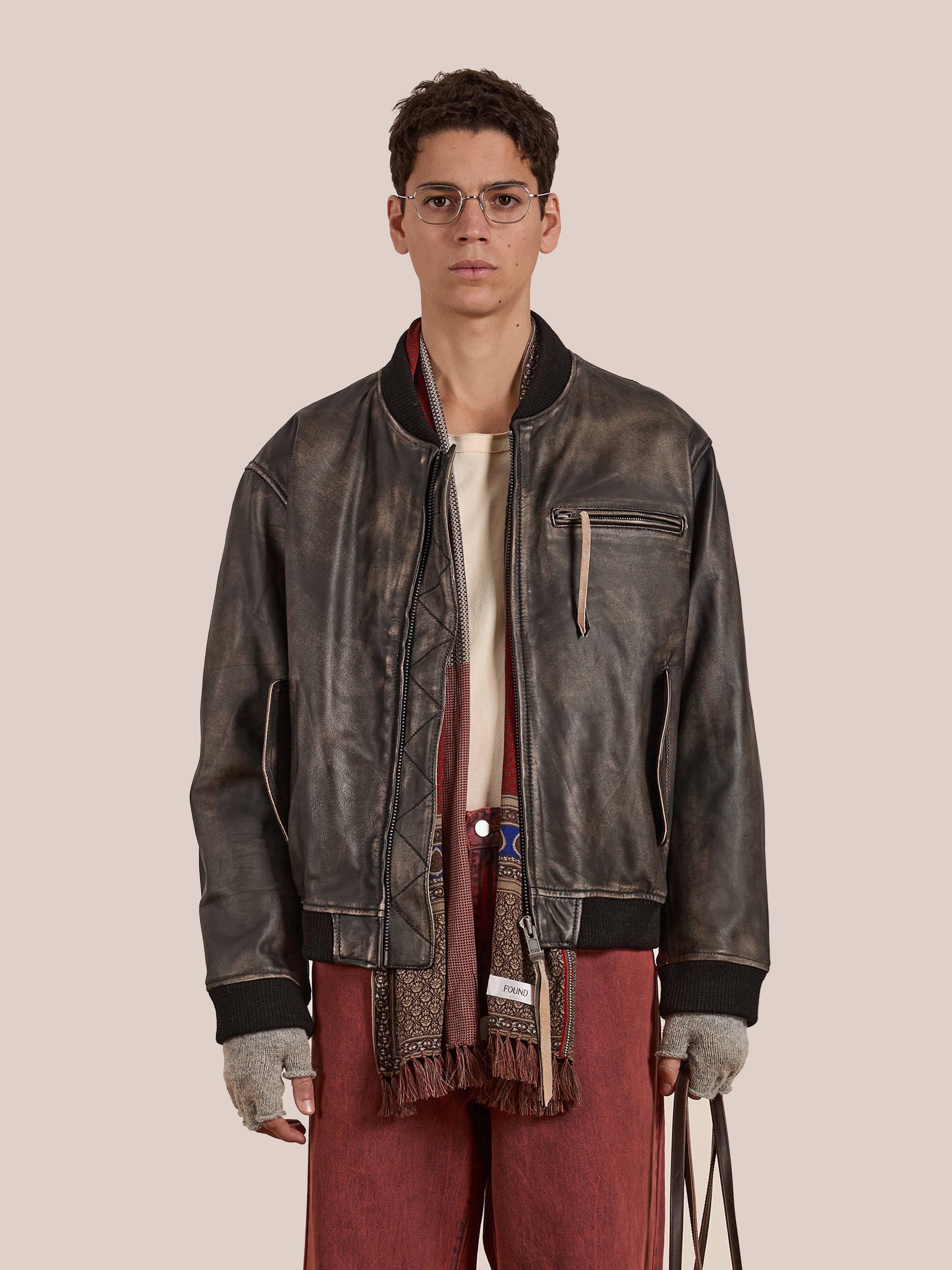 Wearing glasses and long pants, a person holds a bag while dressed in Found's vintage-inspired Distressed Pavement Leather Bomber Jacket made of genuine cowhide leather.