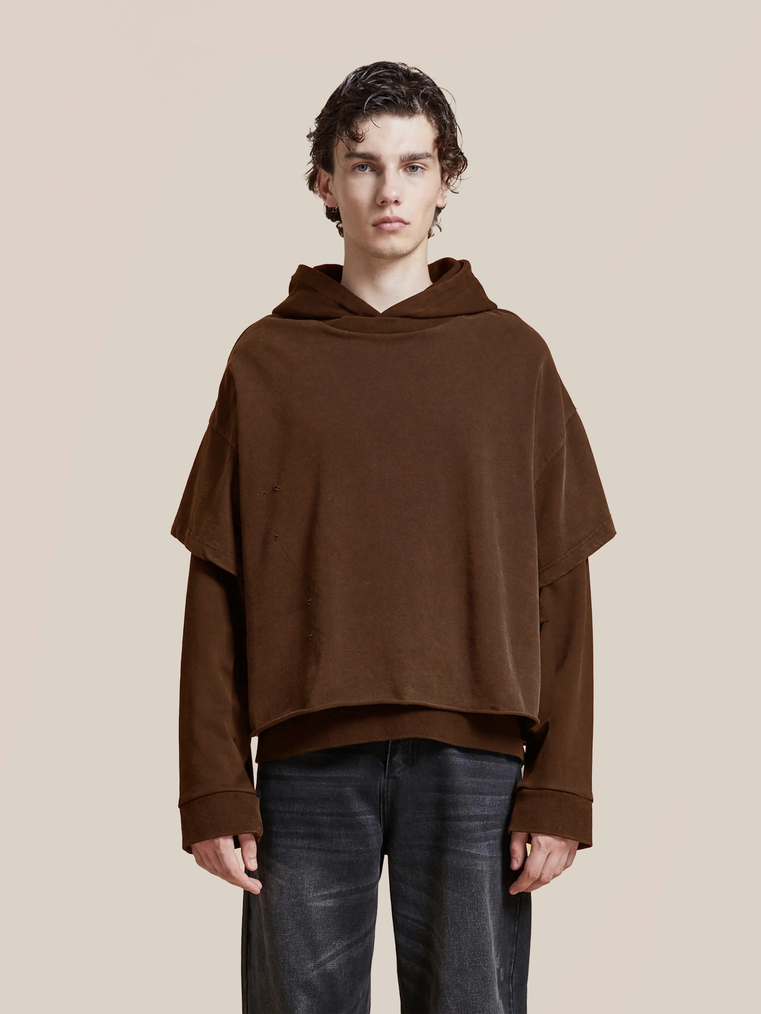 Someone stands against a plain beige background, wearing the Found Double Layer Hoodie—crafted from French terry cotton—in a vintage style, layered over a long-sleeve shirt and paired with dark jeans.