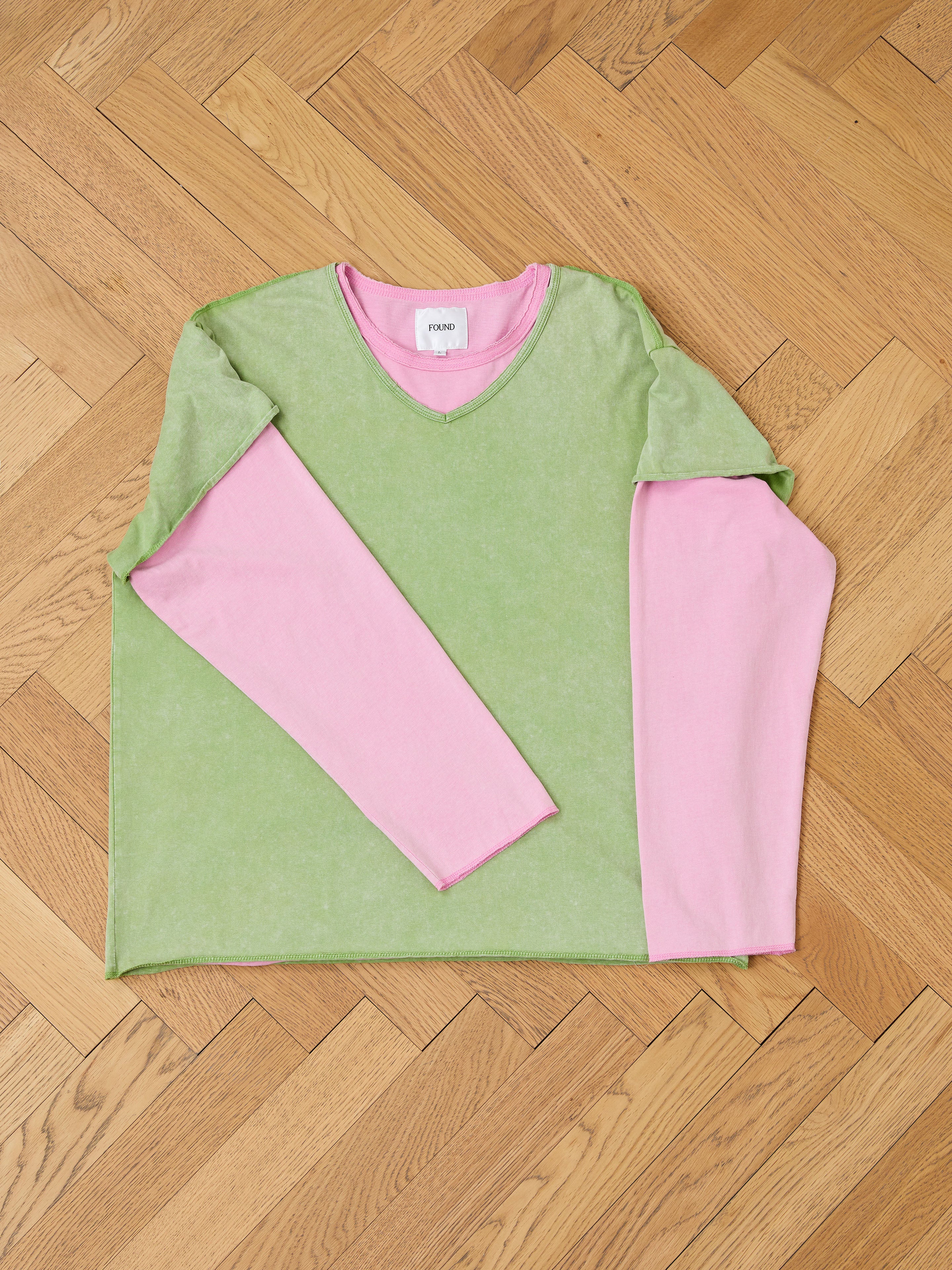 The FOUND Double Layer LS Tee, a unisex long sleeve shirt in green with pink sleeves, is displayed flat on a wooden parquet floor, highlighting its casual style and sunfade wash.