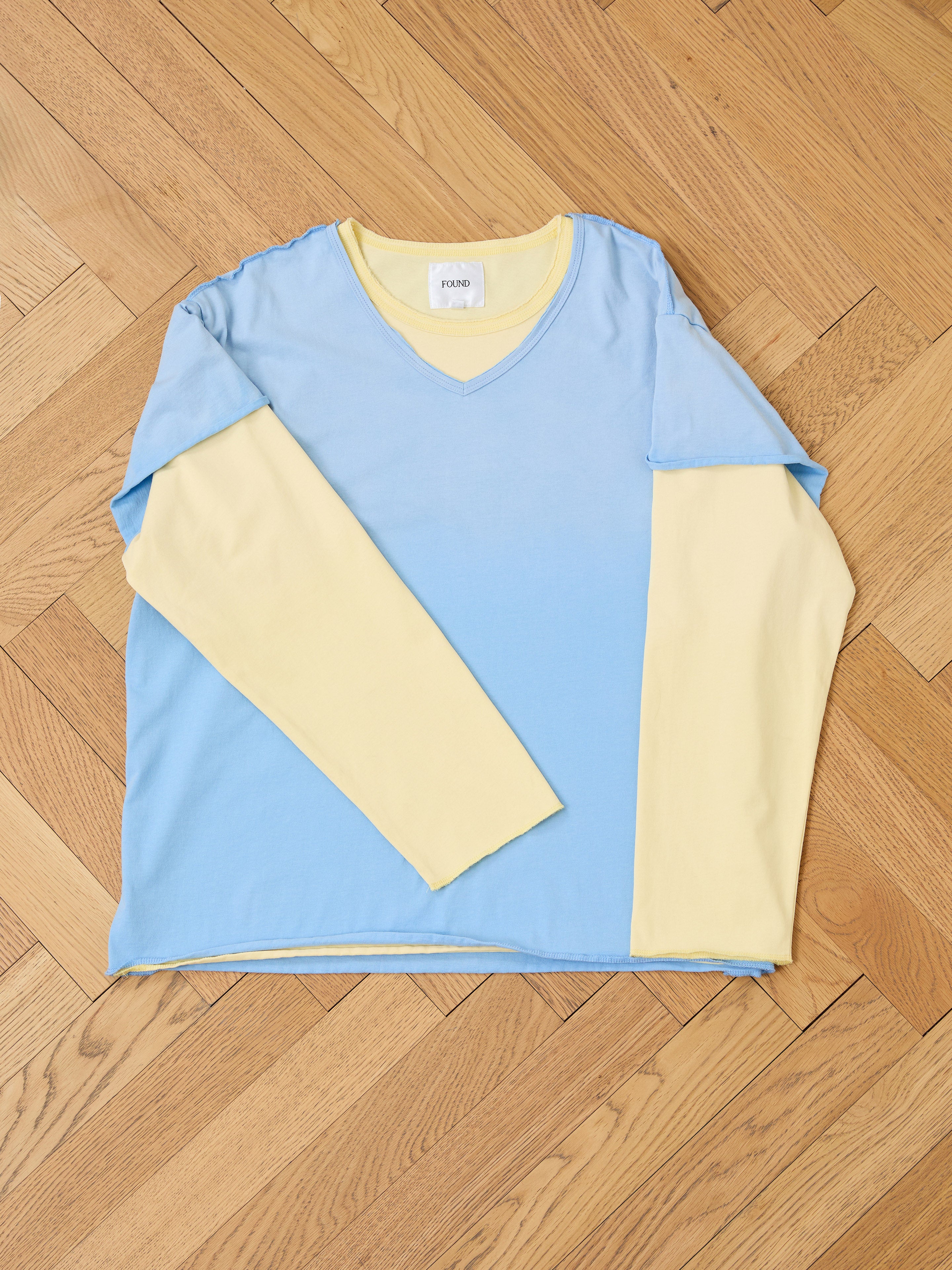 The FOUND Double Layer LS Tee, a unisex long sleeve shirt with a blue sunfade wash body and yellow sleeves, is laid flat on a light wood herringbone floor.