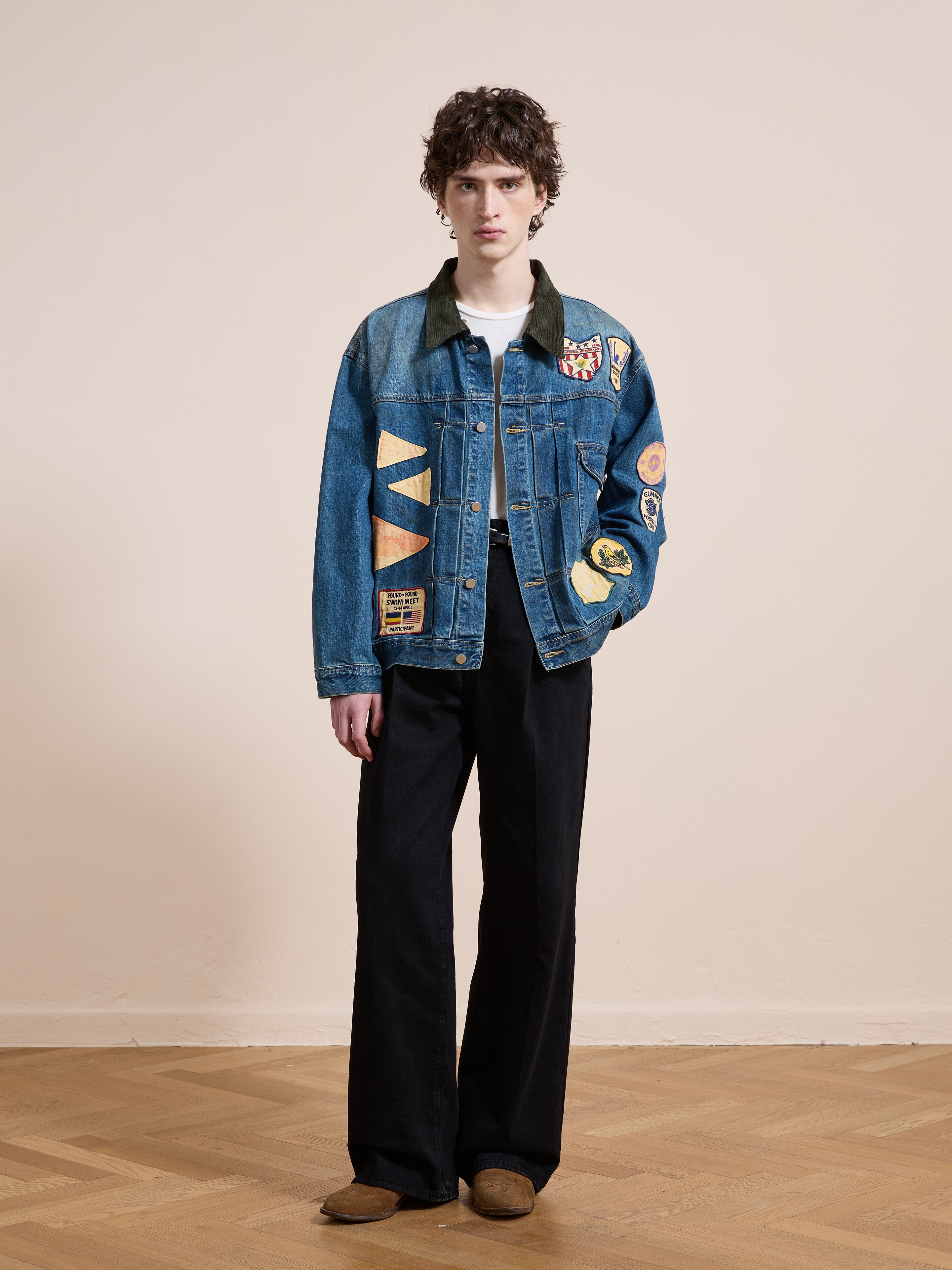 A person stands indoors against a beige wall and wooden floor, wearing the FOUND Denim Patchwork Jacket with various patches, paired with black wide-leg pants and brown shoes.