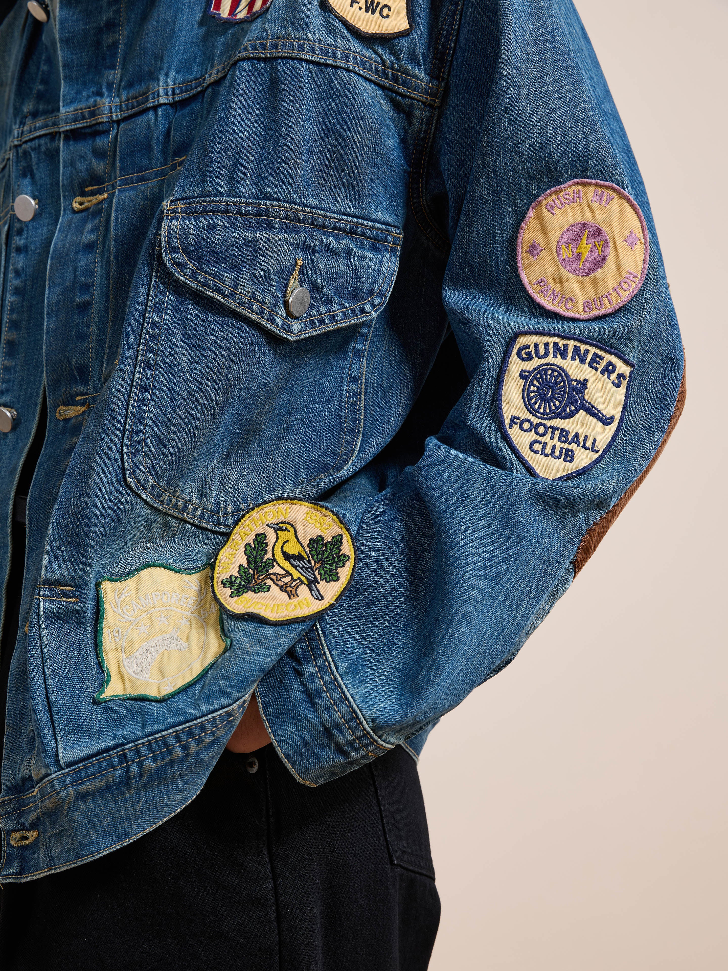 A person sports the FOUND Denim Patchwork Jacket, a unisex relaxed fit with vintage wash and colorful embroidered patches on sleeve and pocket for laid-back charm.