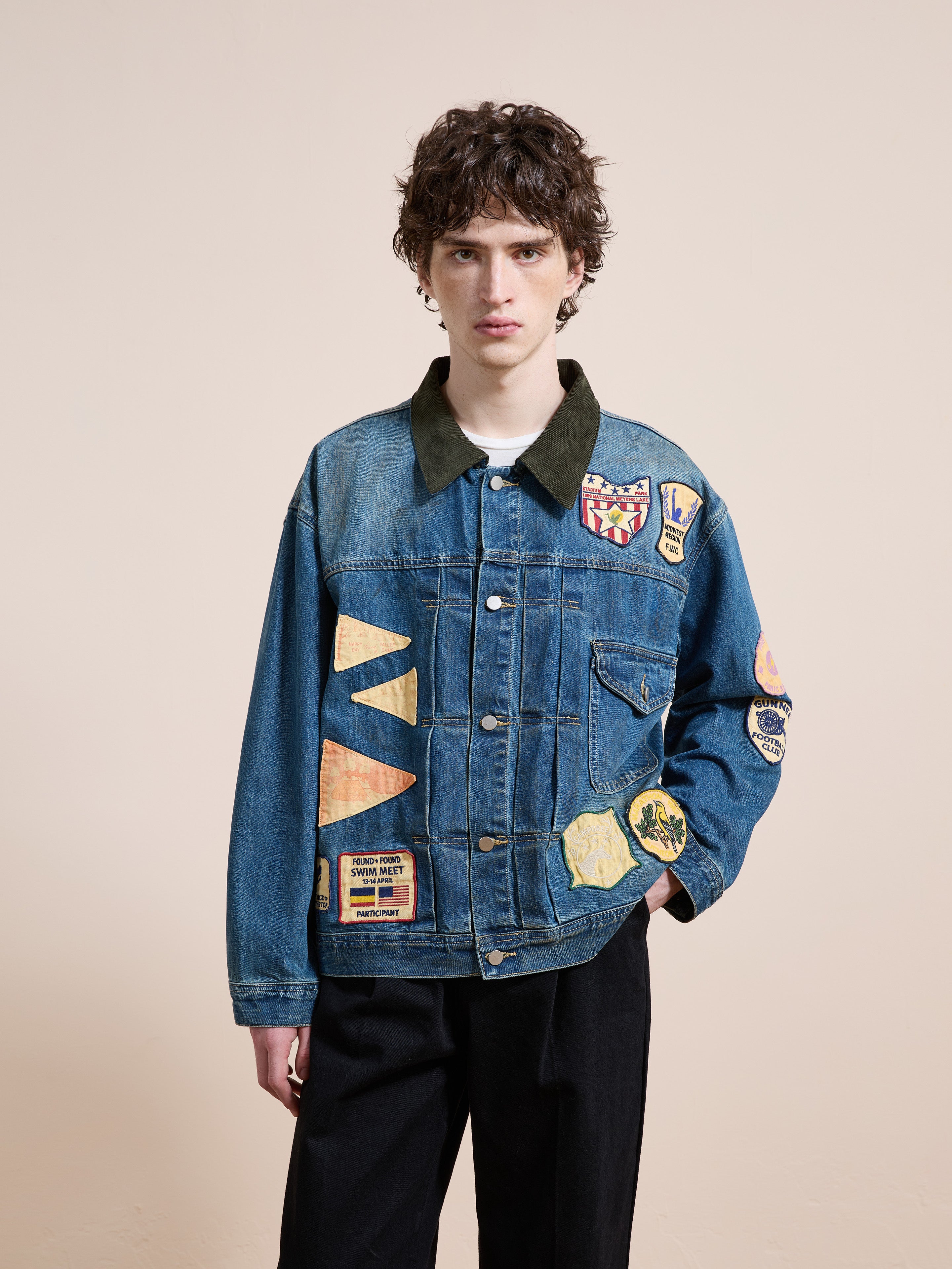 A person stands against a plain background wearing the FOUND Denim Patchwork Jacket, featuring vintage washed denim with colorful patches, paired with black pants.
