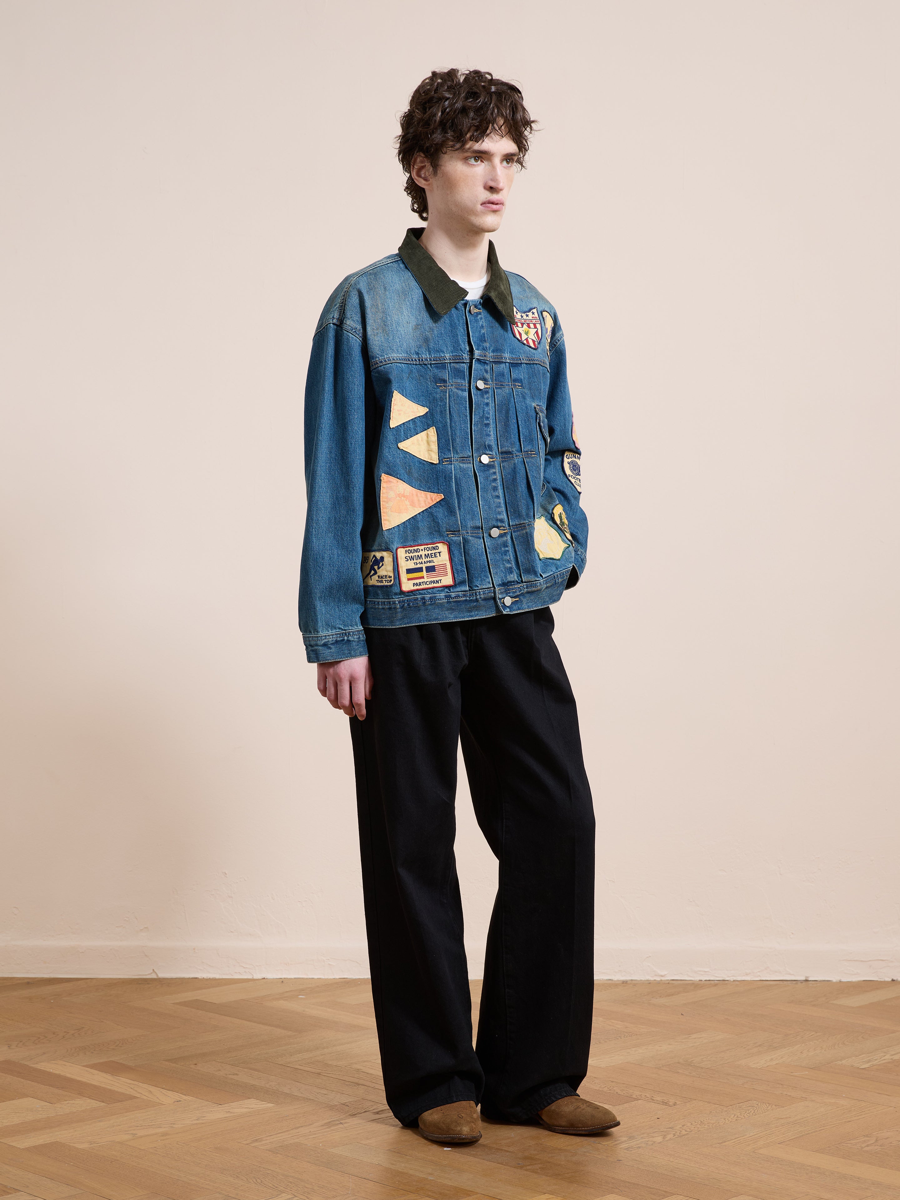 A person stands indoors against a plain background wearing the FOUND Denim Patchwork Jacket, paired with black wide-leg pants and brown shoes.