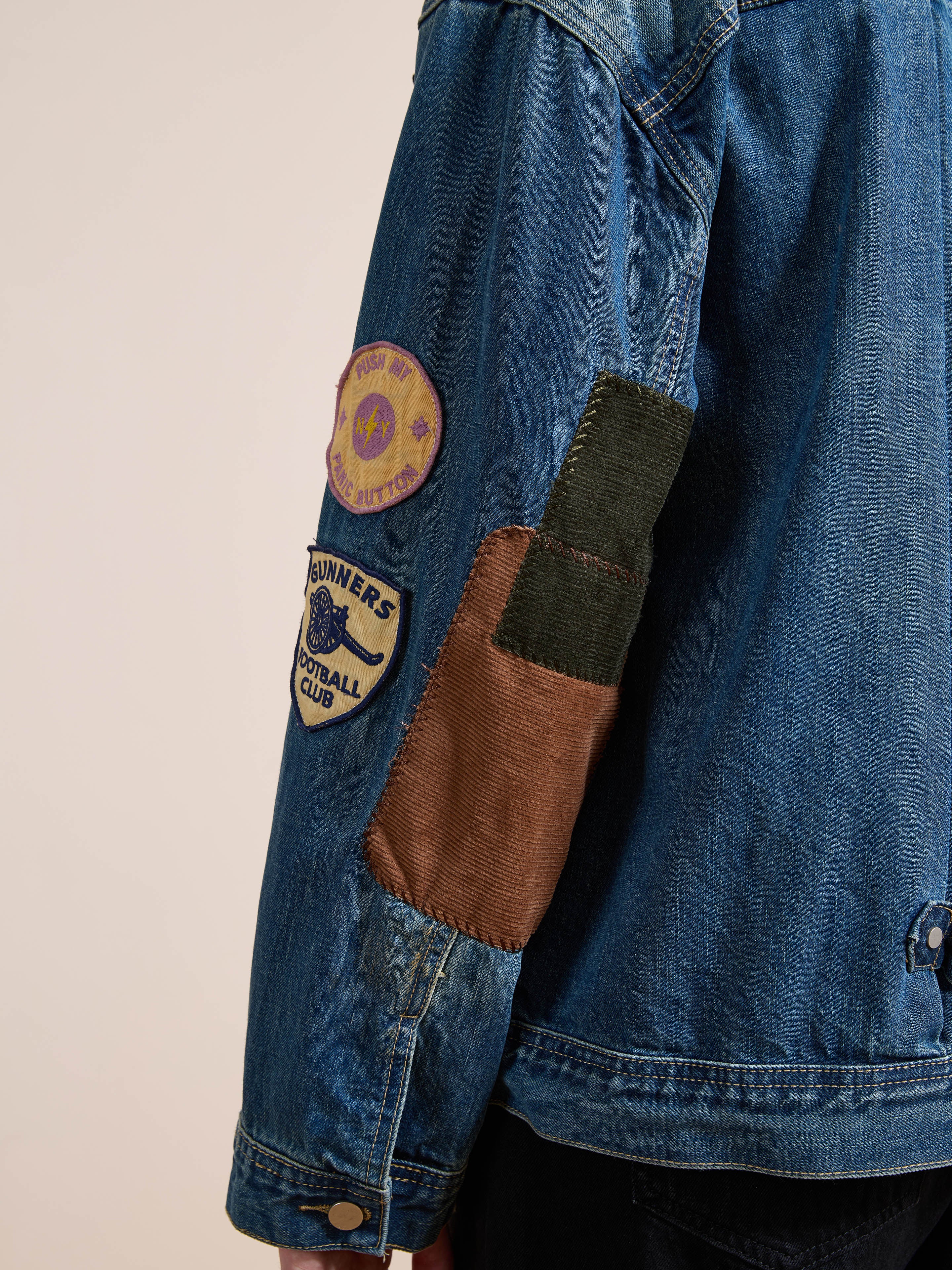 A person photographed from the back wears the FOUND Denim Patchwork Jacket, a vintage-washed, relaxed-fit piece featuring two cloth patches and a multicolored fabric patch on its sleeve.
