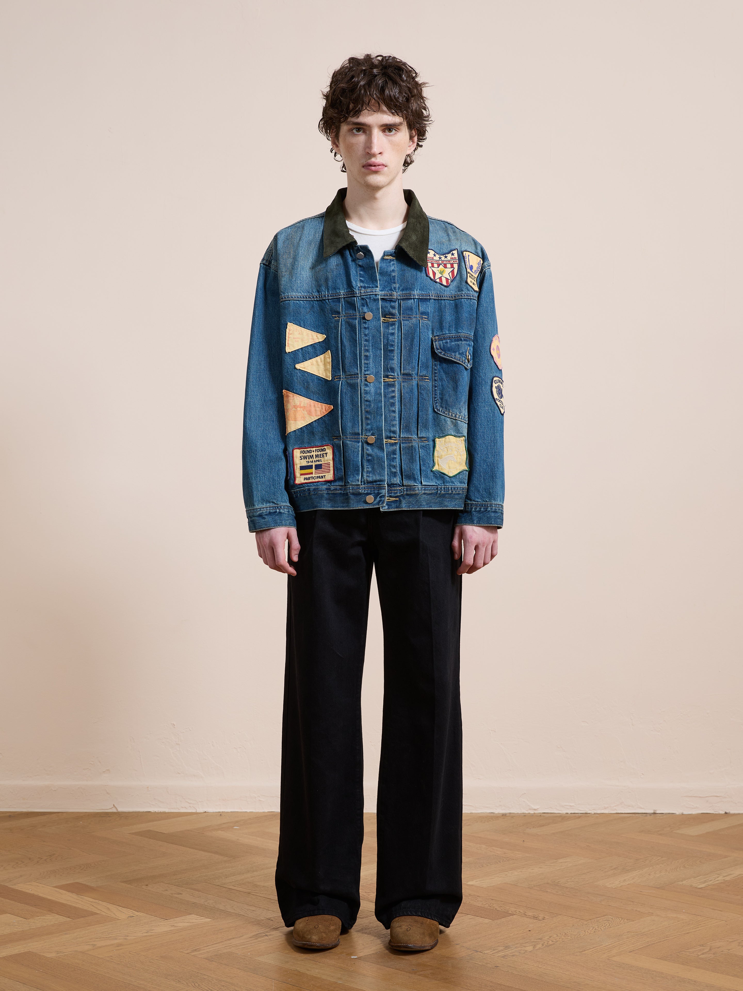 Indoors against a beige wall and wooden floor, a person wears the FOUND Denim Patchwork Jacket with wide-leg black pants and brown shoes.