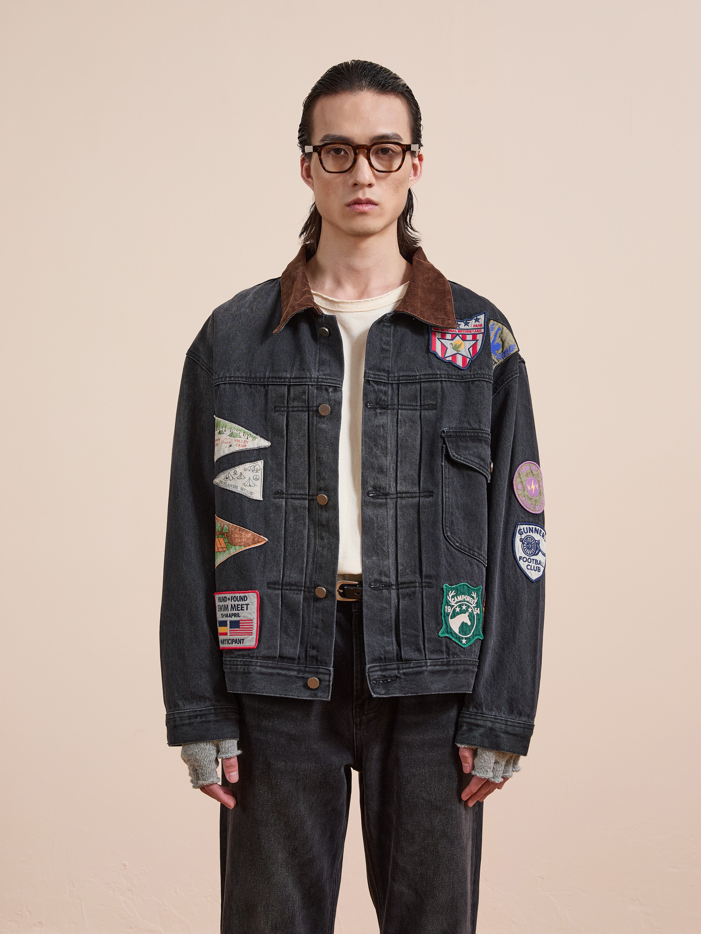 A person with long dark hair and glasses wears the FOUND Denim Patchwork Jacket over a white shirt with fingerless gloves and dark jeans, posing against a plain beige background.