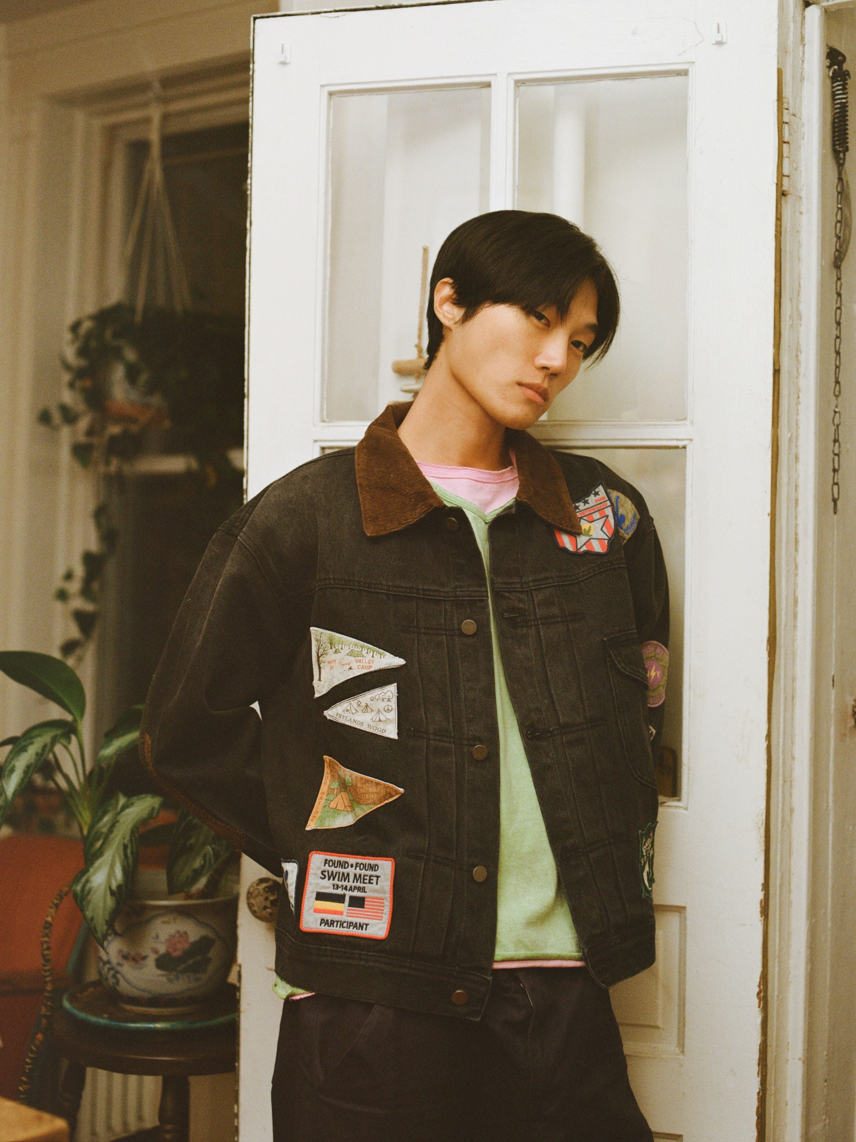 A person with straight black hair stands indoors, wearing the FOUND Denim Patchwork Jacket over a green shirt, with plants and a white door in the background.