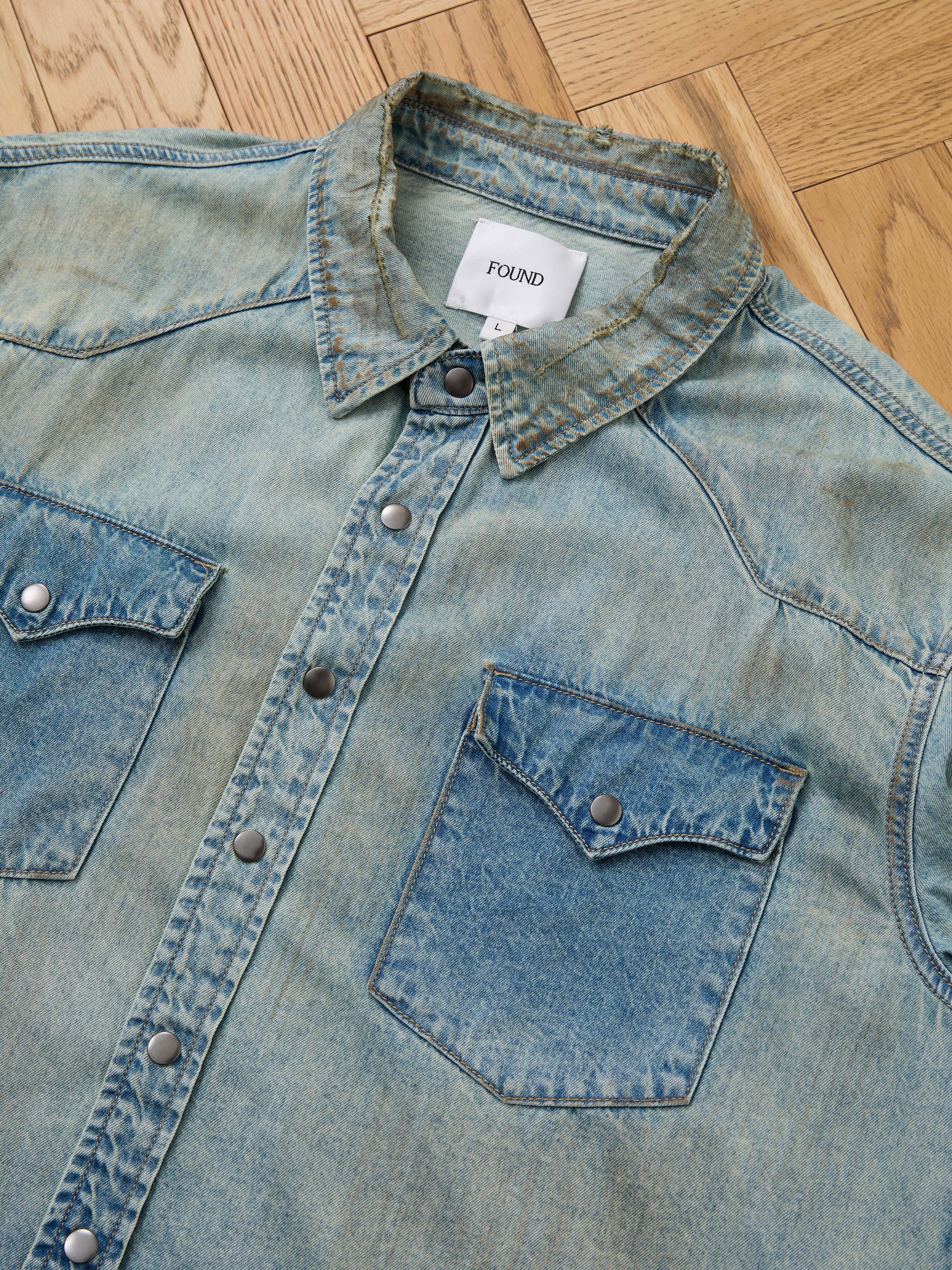 The FOUND Denim Western Snap Button Shirt features a light blue vintage wash, western yoke detailing, snap buttons, and two chest pockets, laid flat on a wooden floor.