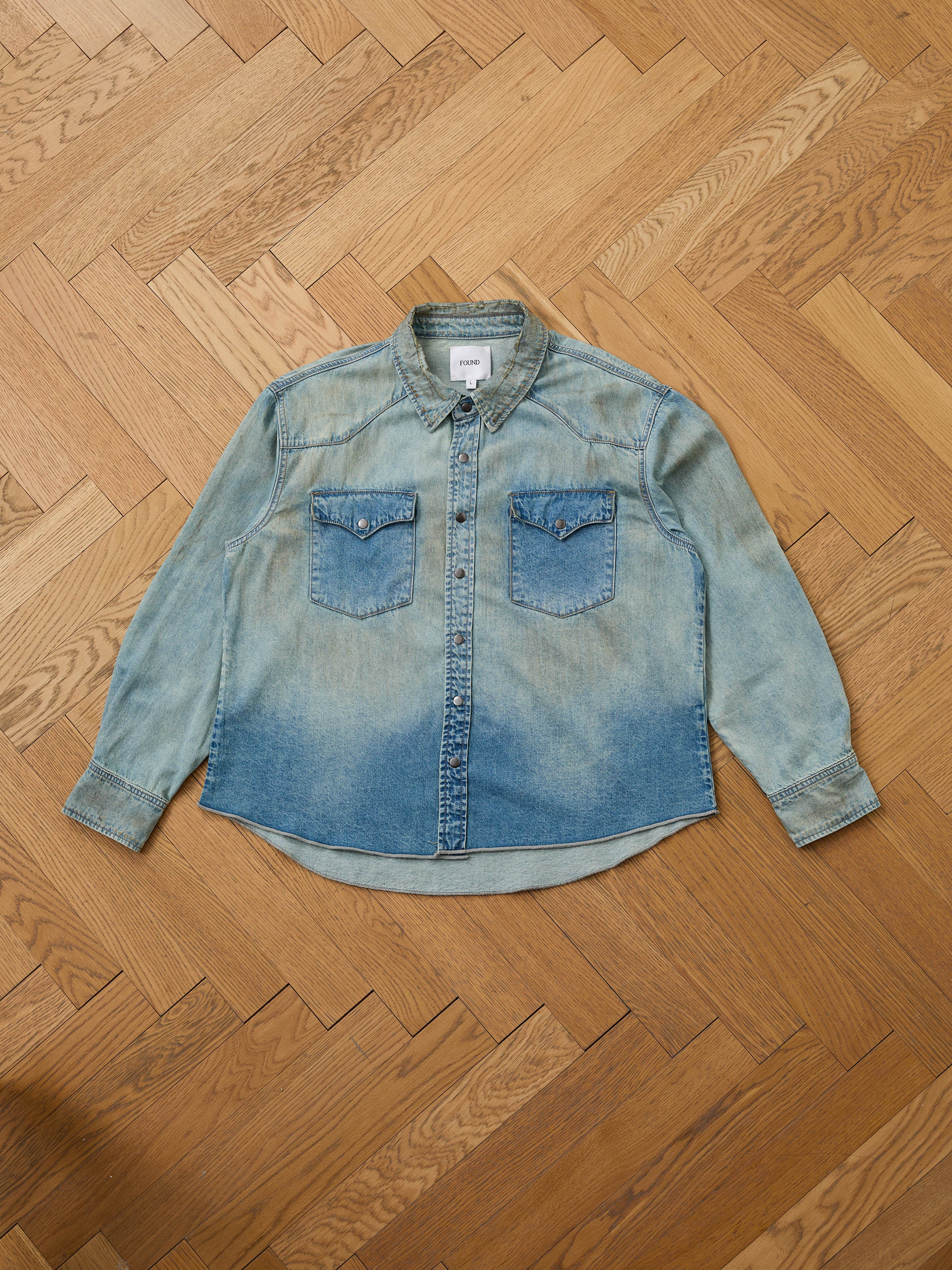 The FOUND Denim Western Snap Button Shirt, in light blue vintage-wash with two chest pockets and western yoke details, is displayed flat on a wooden herringbone-patterned floor.