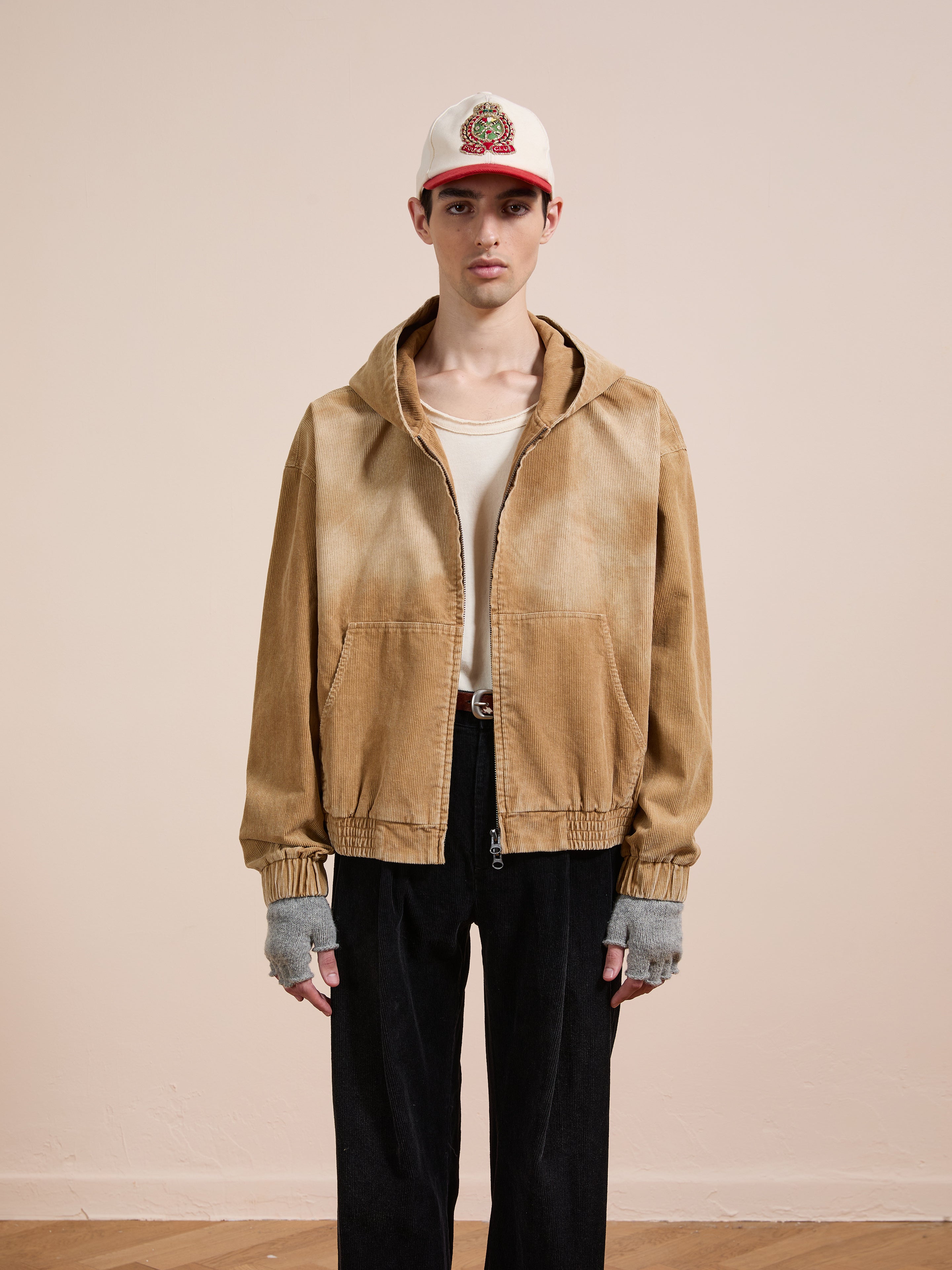 A person stands against a plain background wearing the FOUND Corduroy Zip Up Hoodie in beige, styled with a white shirt, black pants, fingerless gloves, and a white baseball cap featuring a red brim and embroidered patch for a vintage look.