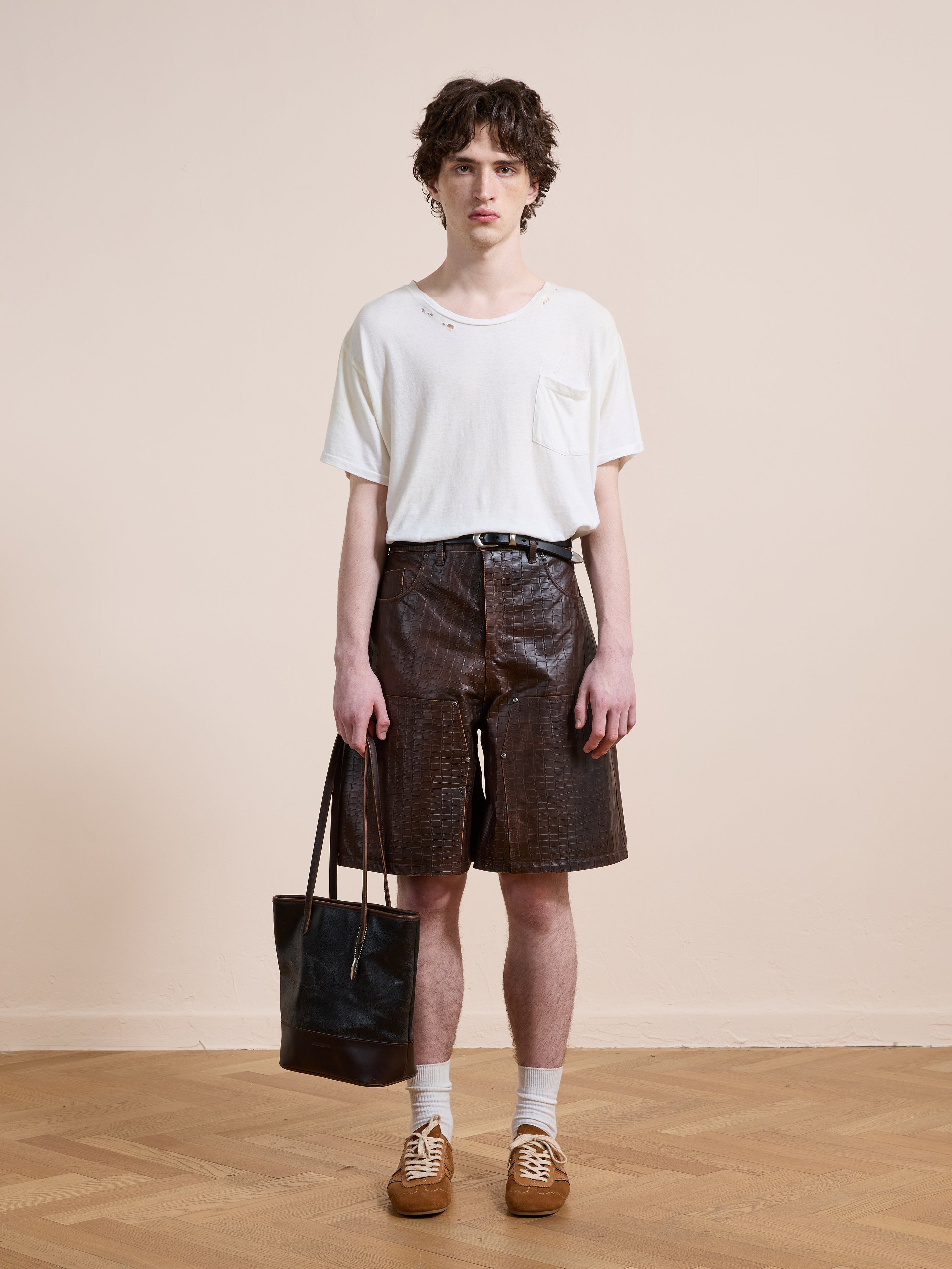 Person stands indoors against a beige wall, wearing a white t-shirt, FOUND Distressed Croc Embossed Leather Shorts in brown, tan sneakers, white socks, and holding a black tote bag.