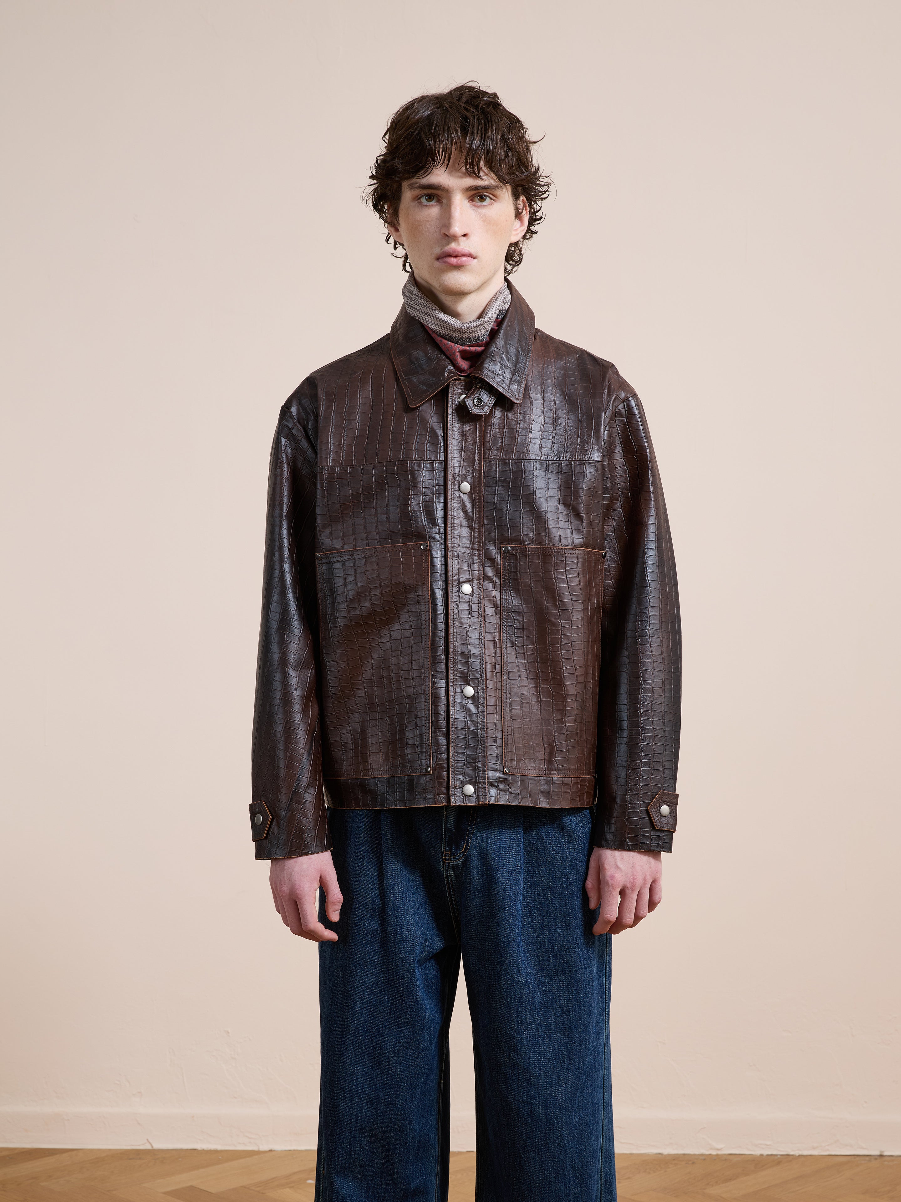 A person in the FOUND Distressed Croc Embossed Leather Work Jacket, layered over a turtleneck and paired with wide-leg blue jeans, stands against a plain beige wall.