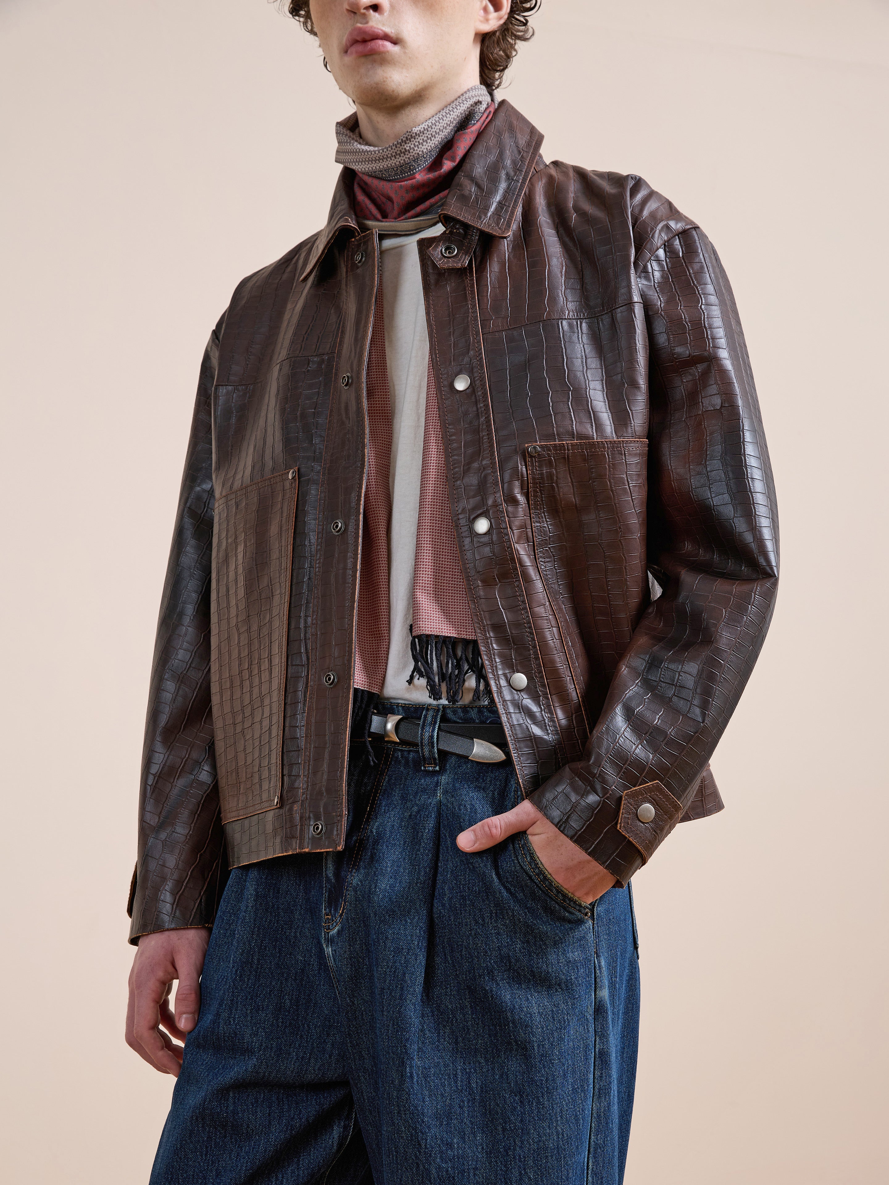A person wears the FOUND Distressed Croc Embossed Leather Work Jacket over layered shirts and blue jeans, standing with one hand in their pocket against a neutral background.