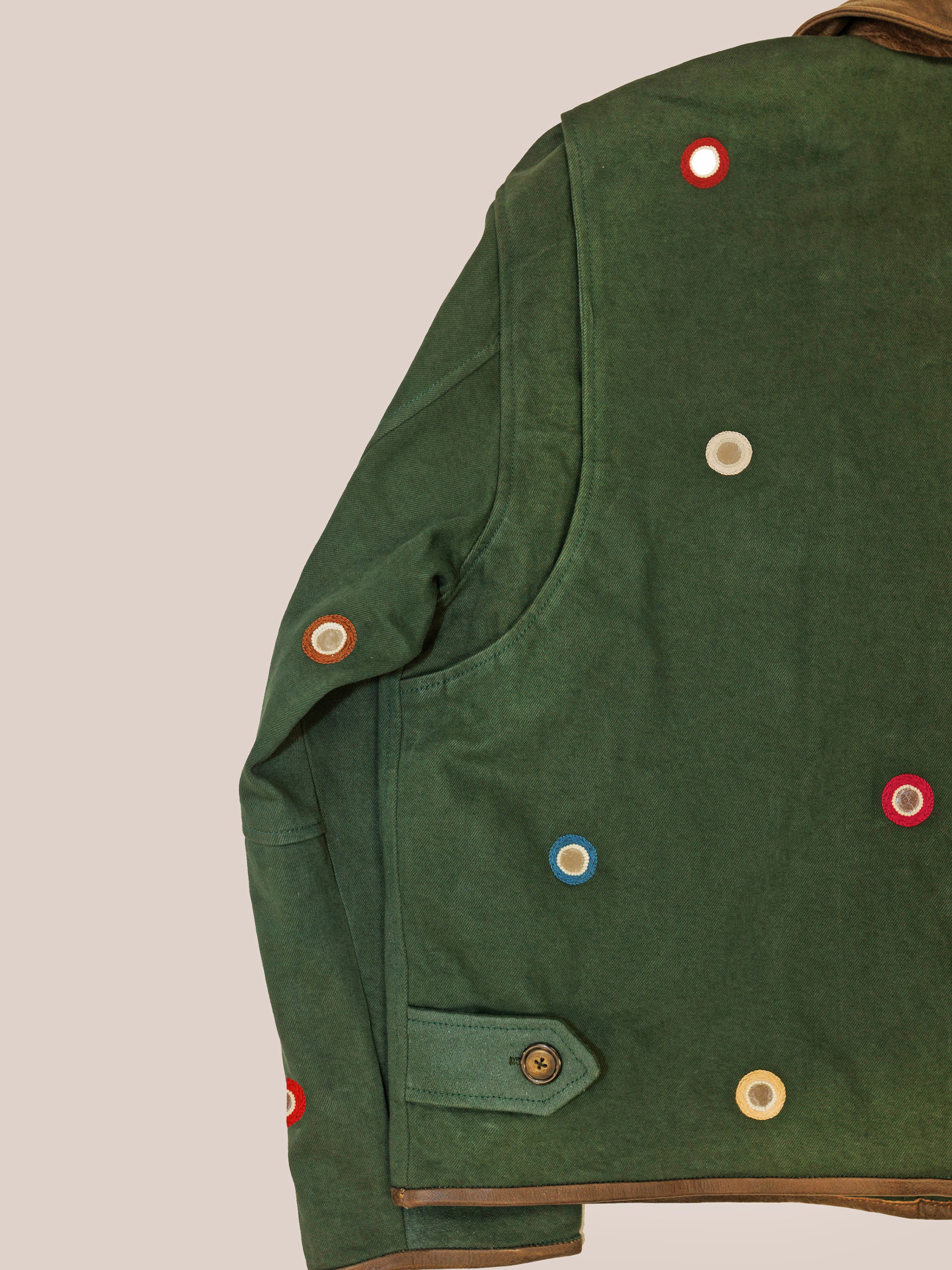 The Hand Embroidered Mirror Abhala Jacket by Found, shown from the back on a plain beige background, features green cotton twill and multicolored circular patches with a buttoned pocket.
