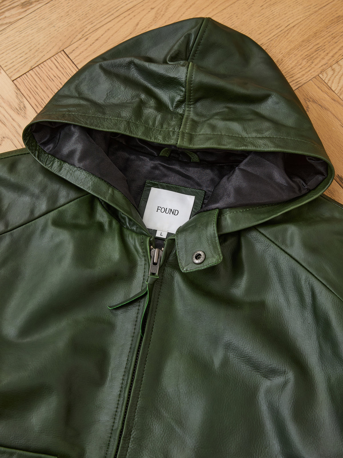 A green Distressed Leather Pocket Hoodie by FOUND, featuring a zipper and displayed on a wooden floor. The inner tag reads “FOUND L.”.