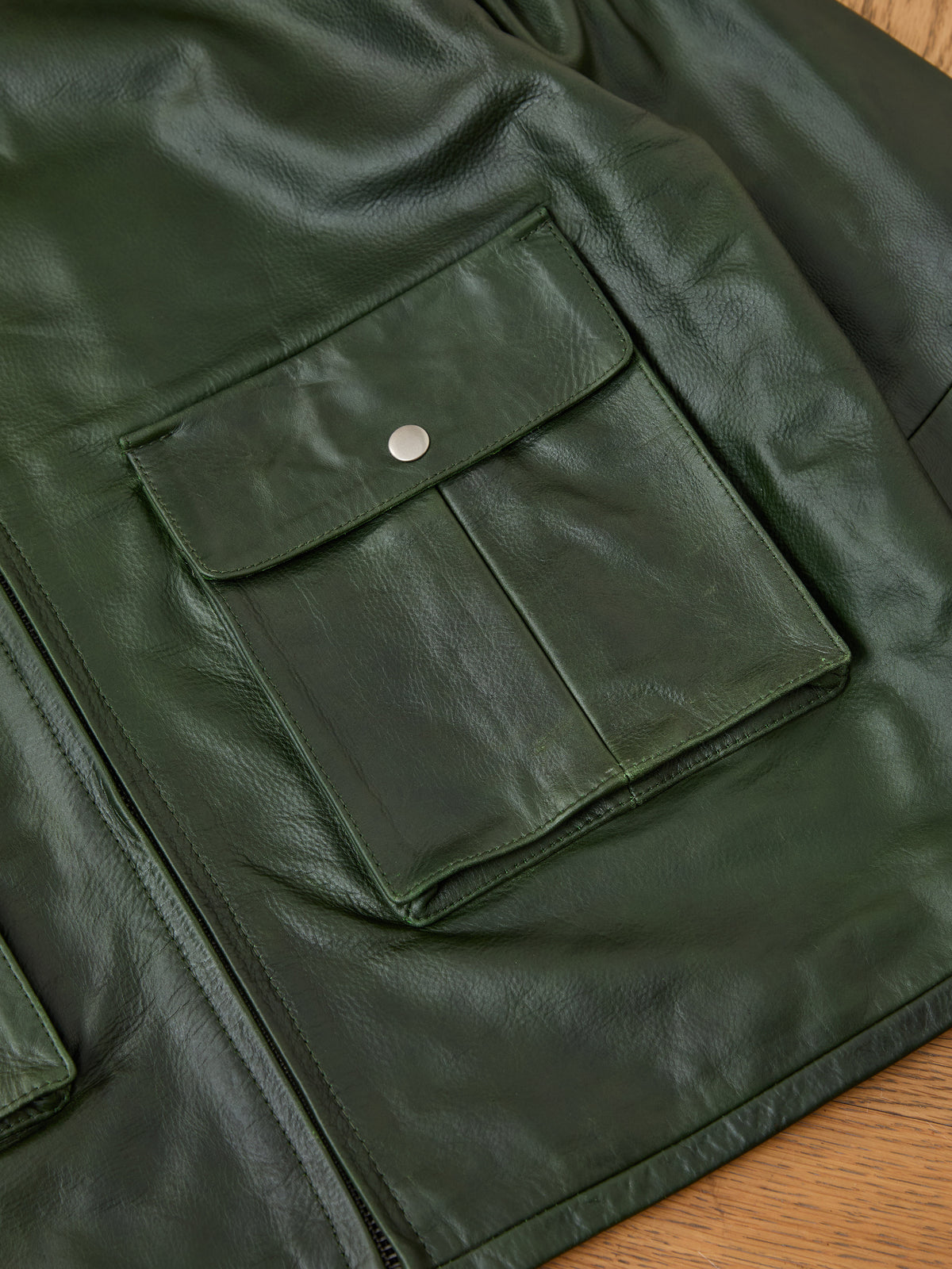 Close-up of the FOUND Distressed Leather Pocket Hoodie in green leather, showcasing a front pocket with a flap and silver snap button, laid out on a wooden surface—a stylish essential blending classic jacket details with hoodie-inspired design.