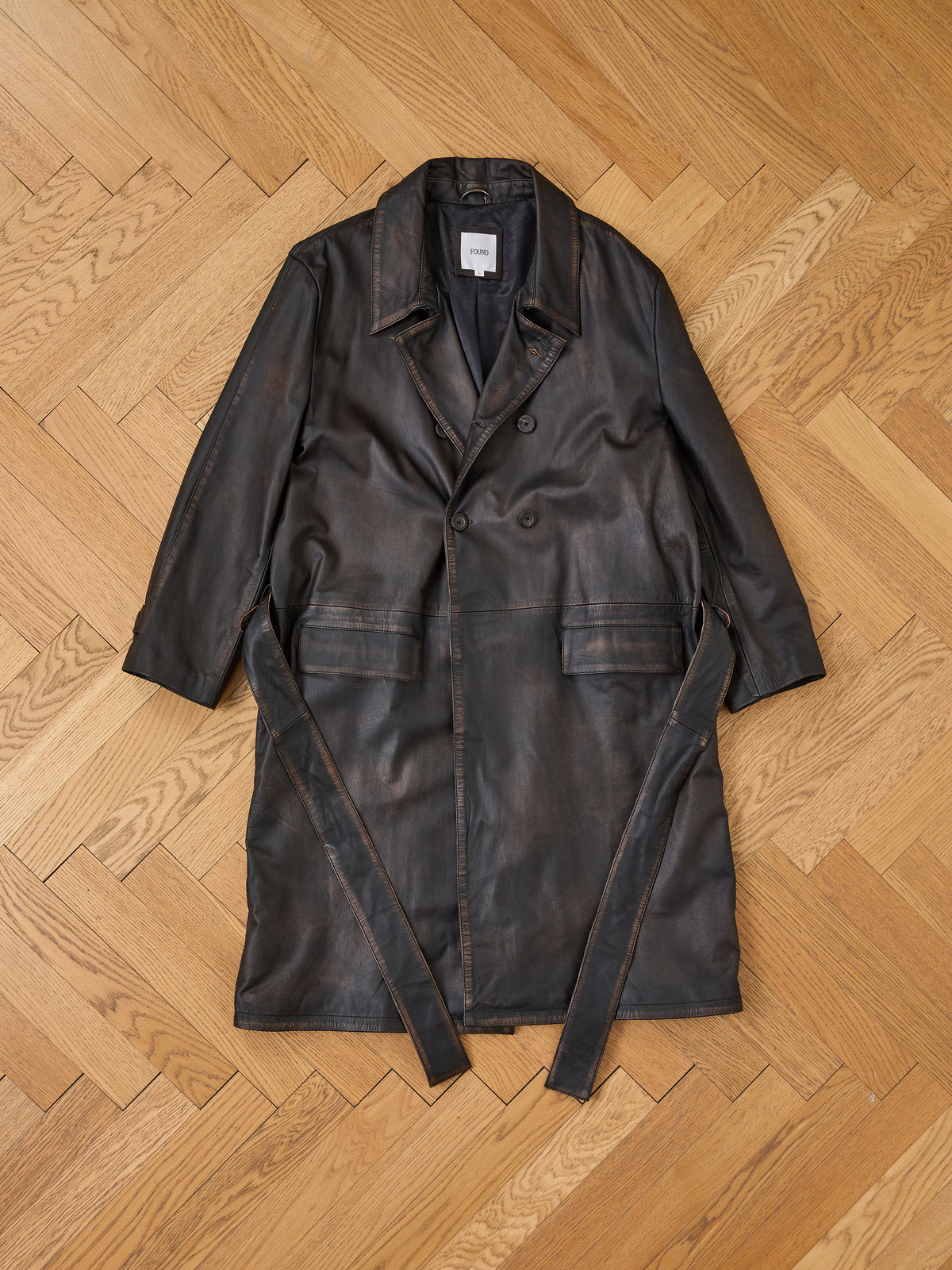 The FOUND Belted Leather Trench Coat, ethically crafted for a unisex fit, is laid flat on a light wood herringbone floor.
