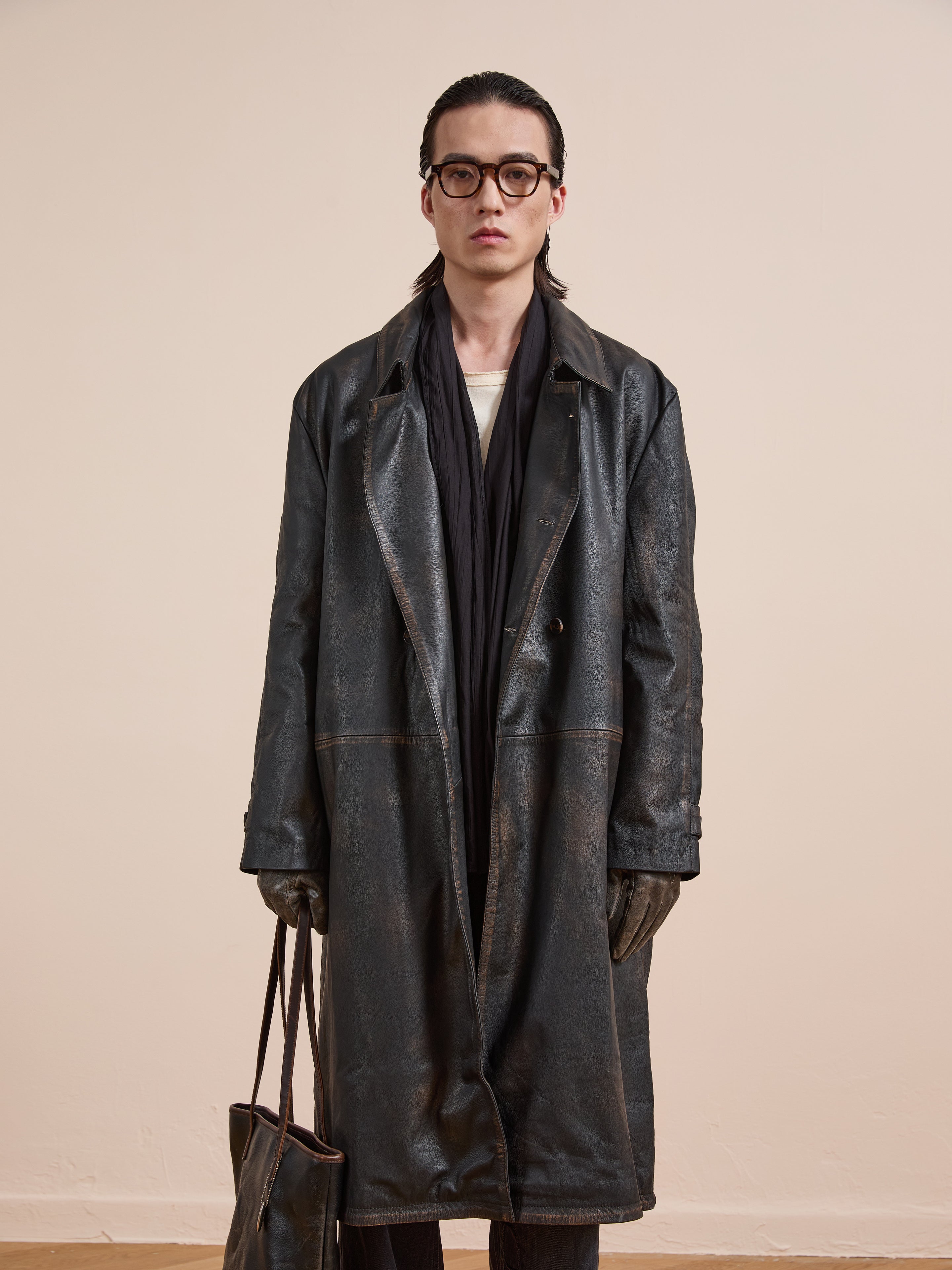 A person in FOUND’s Distressed Leather Trench Coat stands facing the camera with a matching bag and neutral expression, set against a plain background.