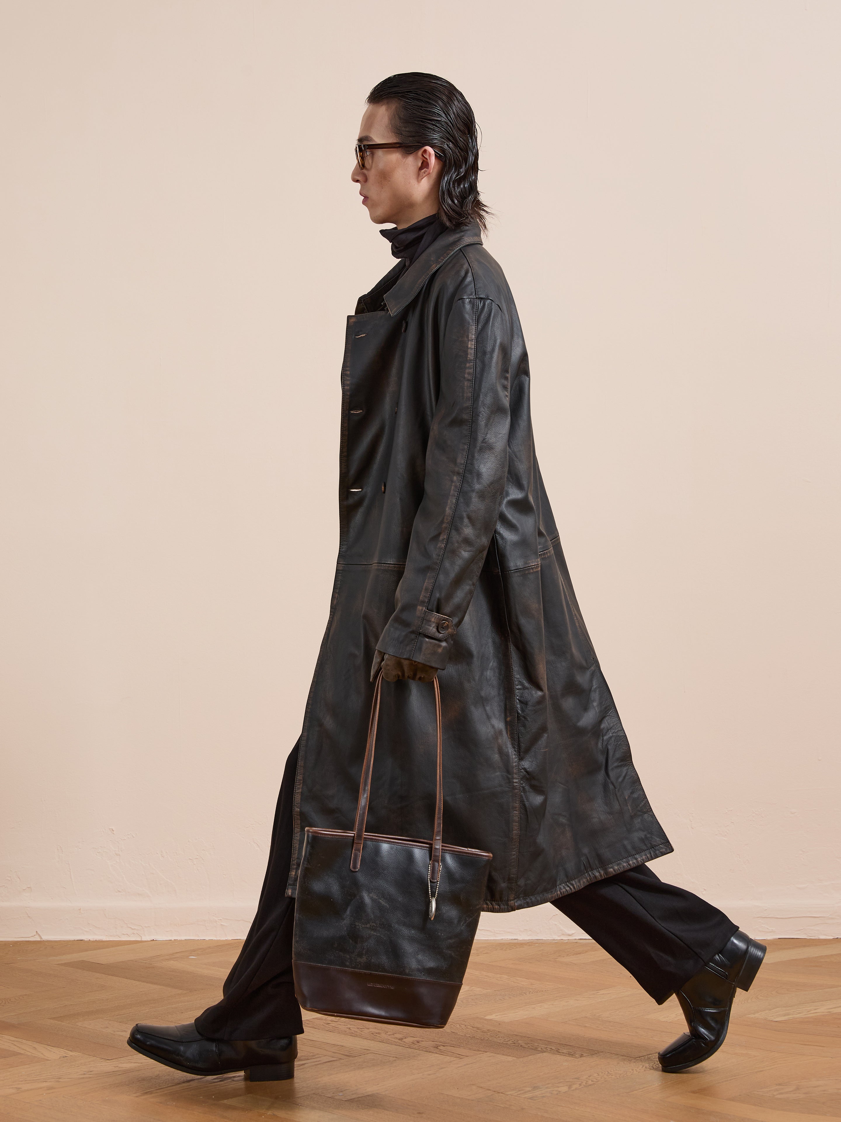 A person walks indoors in a FOUND Distressed Leather Trench Coat, black pants, and loafers, carrying a black and brown tote bag against a plain beige background.