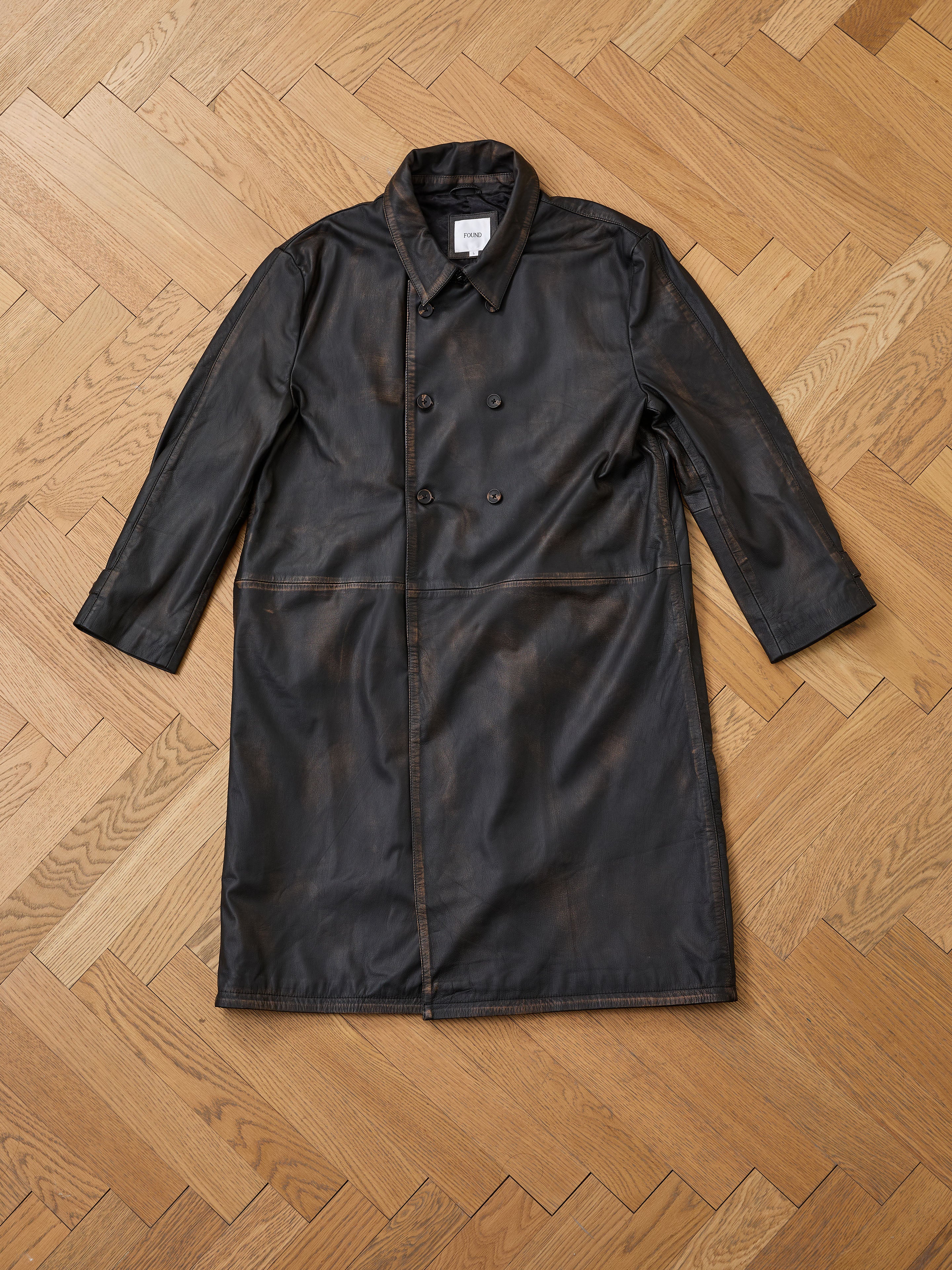 The FOUND Distressed Leather Trench Coat, a long double-breasted genuine leather style with a collar, is displayed flat on a wooden herringbone floor.